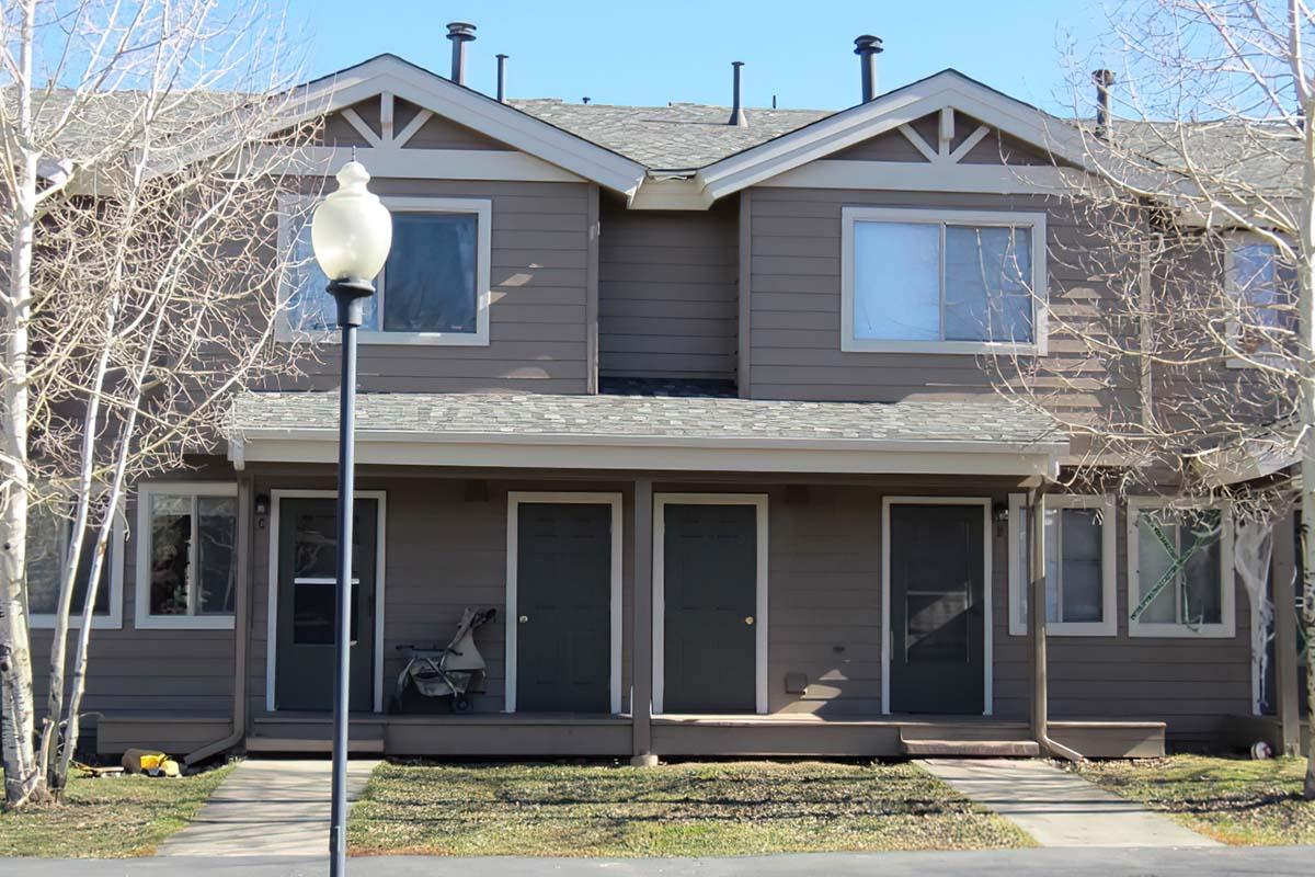A two-story townhouse with a gray exterior, featuring two front doors and large windows. The building is flanked by bare trees, and there’s a lamppost in the foreground. The path leading to the doors is made of wooden planks, and the area is landscaped with grass.