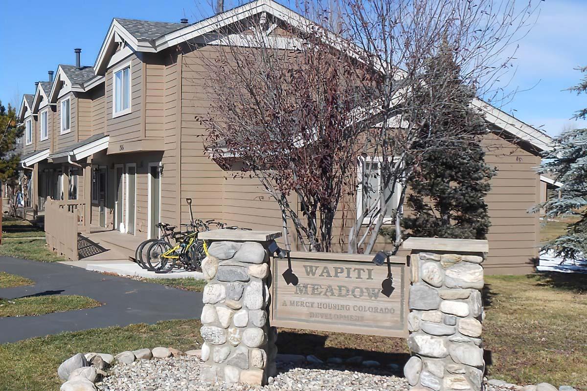 A two-story brown house in a residential area with a sign that reads "Wapiti Meadow" in front. The house has a porch, and two bicycles are parked nearby. Trees and shrubs are present in the yard, and the sky is clear with a few clouds. The scene depicts a quiet, suburban setting in winter.