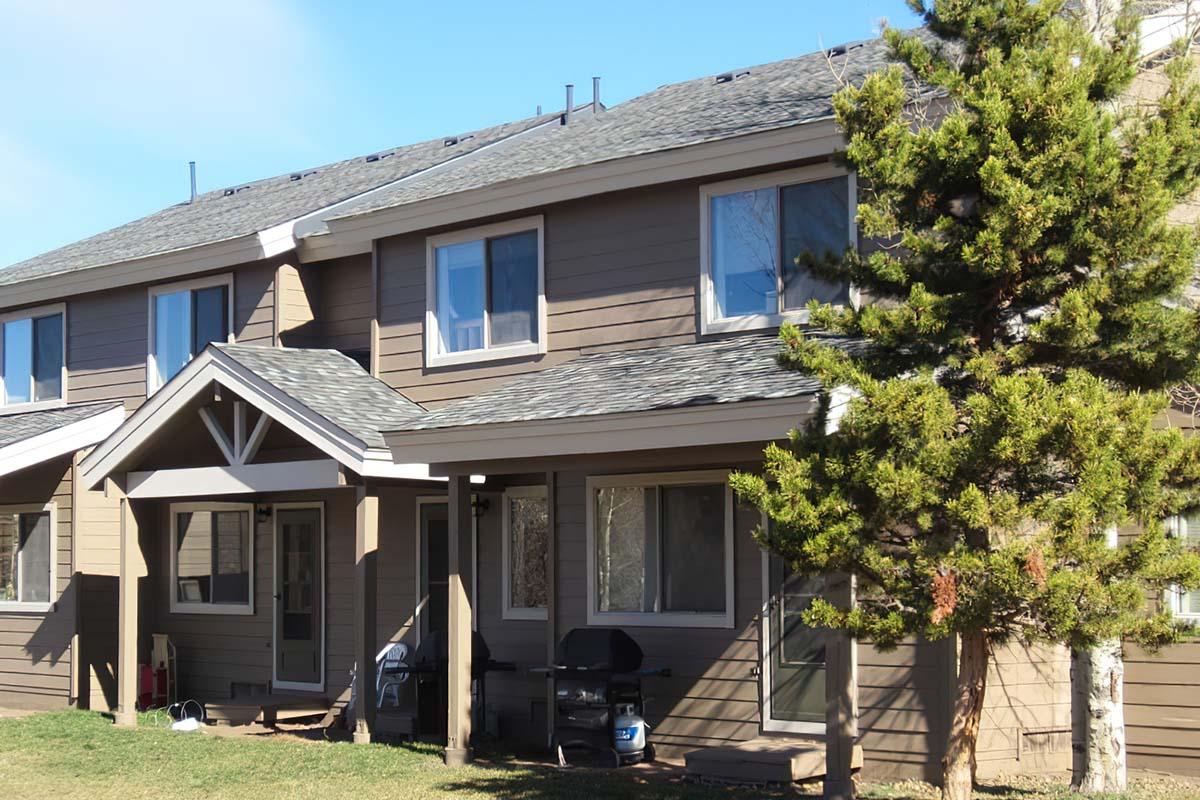 Two-story brown house with a sloped roof, featuring multiple windows and a covered porch. A green pine tree stands in the foreground, and there is outdoor furniture on the porch. The lawn is well-maintained, and the sky is partly cloudy.