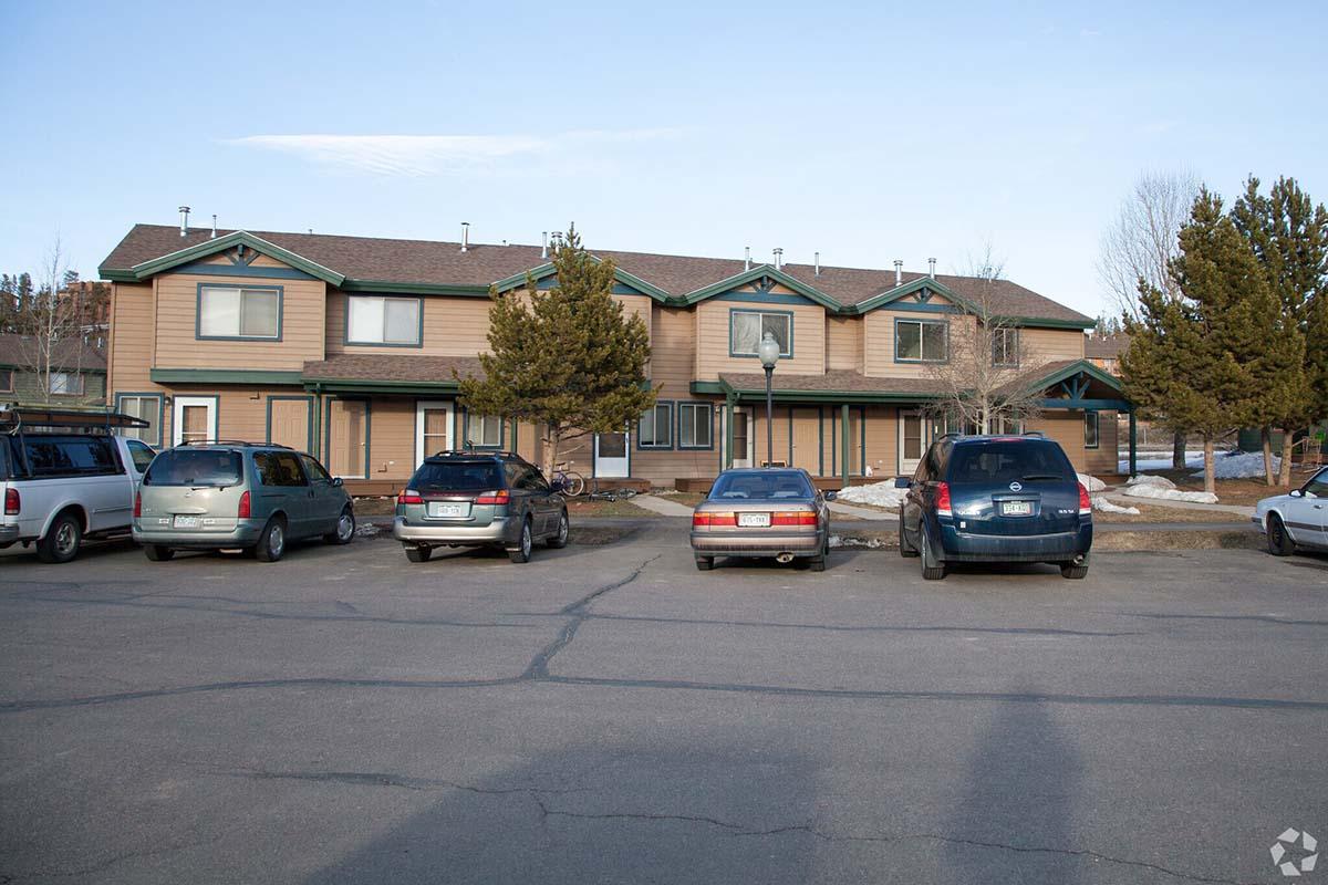 A row of residential townhouses with brown exterior and green accents, set against a clear blue sky. Several parked cars are visible in the foreground, with patches of snow on the ground, indicating early spring or late winter. Trees are present nearby, adding to the suburban atmosphere.