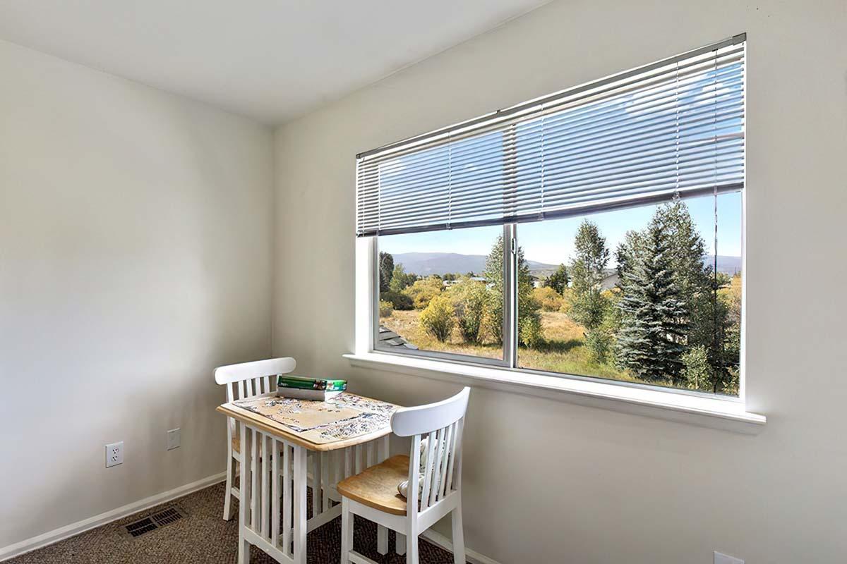 A bright, minimalist room featuring a small table with two white chairs, situated near a window. Outside, a scenic view of trees and grasslands under a clear sky can be seen. The interior walls are painted a light color, enhancing the sense of space and light.