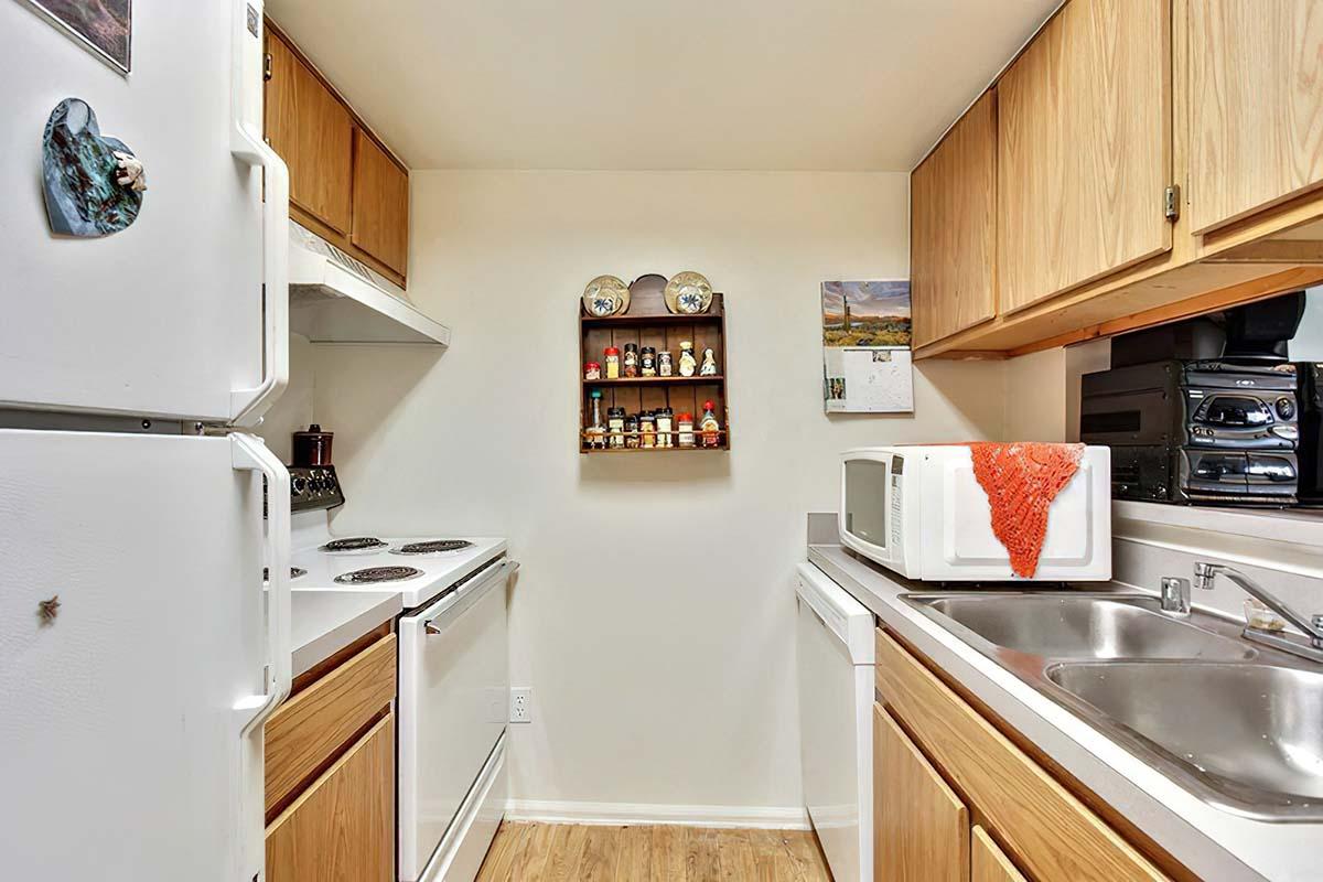 A small kitchen featuring white appliances including a refrigerator, stove, and microwave. The wooden cabinets above and below provide storage, while a spice rack and a calendar adorn the wall. A double sink is visible, and the floor is made of light-colored wood.