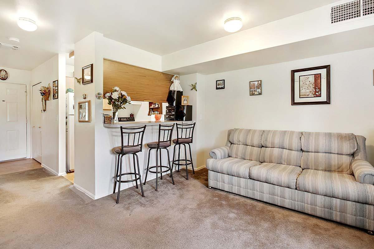 A cozy living area featuring a striped sofa, a small bar with stools, and framed pictures on the walls. The space has neutral-colored walls and carpeting, with soft lighting and decorative items like plants and photo frames. The entrance leads to a kitchen area visible in the background.