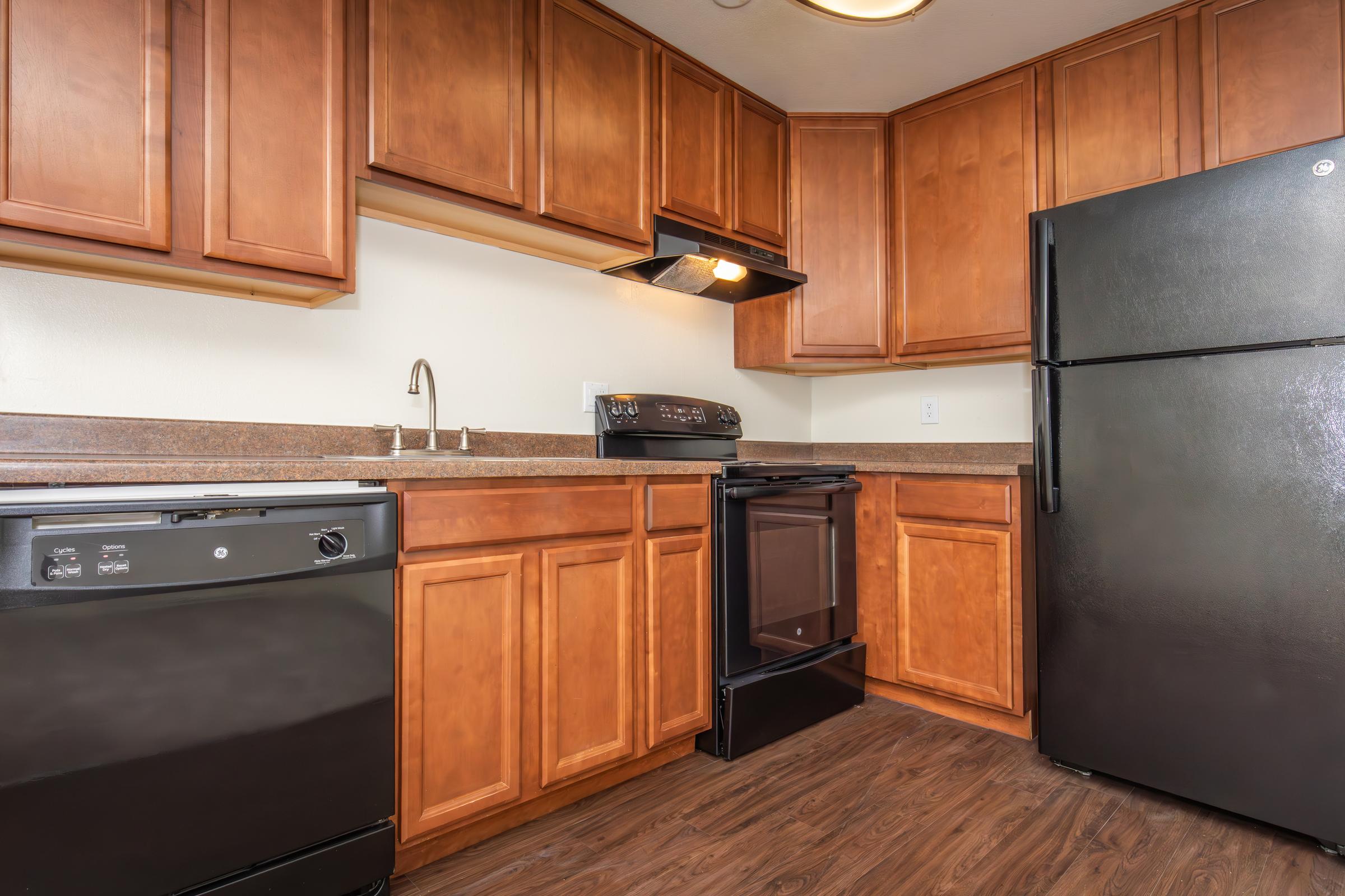 A small kitchen featuring wooden cabinets, a black refrigerator, a stainless steel gas stove with an oven, and a countertop with a sink. The walls are a light color, and the flooring is dark wood. No visible kitchen appliances other than the stove and refrigerator.