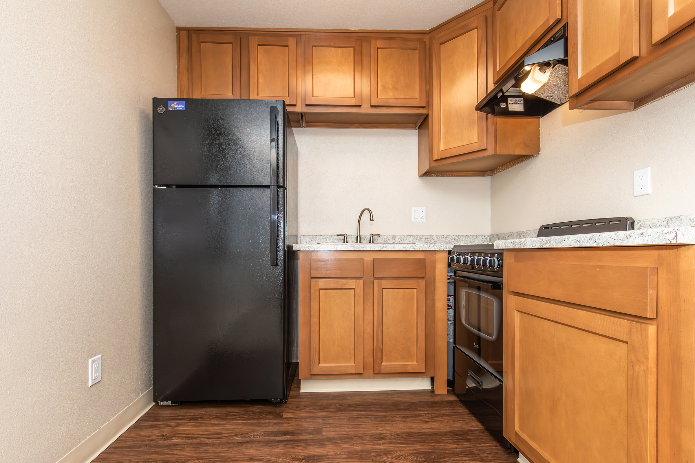 A modern kitchen featuring wooden cabinetry, a black refrigerator, a gas stove, and a sink. The countertops are light-colored with a speckled pattern. The floor is dark wood, and the walls are painted in a neutral tone. The space is well-lit, providing a cozy atmosphere.