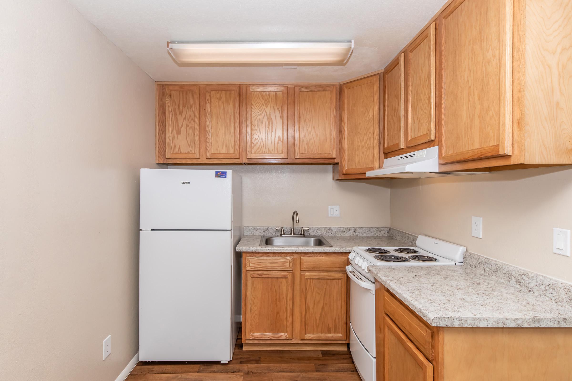 A small kitchen featuring wooden cabinets, a white refrigerator, a sink, and a stove with an oven. The countertops are light-colored, and the walls are painted beige, creating a warm and inviting atmosphere. A ceiling light provides illumination in the space.