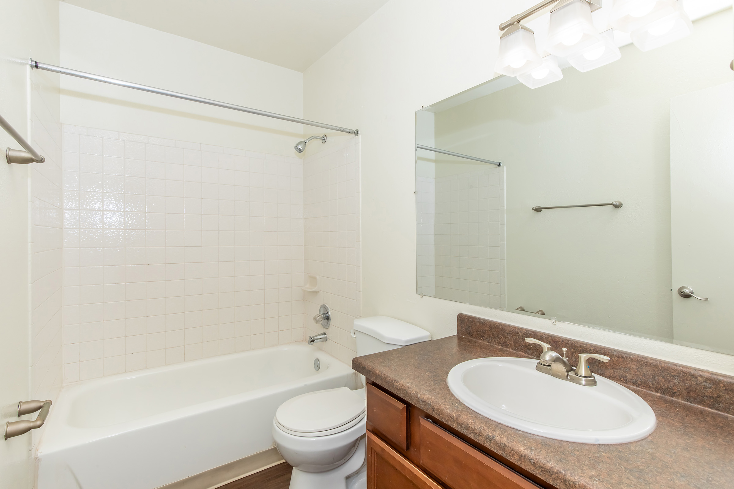 A clean, simple bathroom featuring a white bathtub with a shower, a toilet, and a single sink with a round mirror above it. The walls are painted light-colored, and there are overhead lights above the sink area. The flooring is a warm wood tone, creating a neutral and inviting atmosphere.