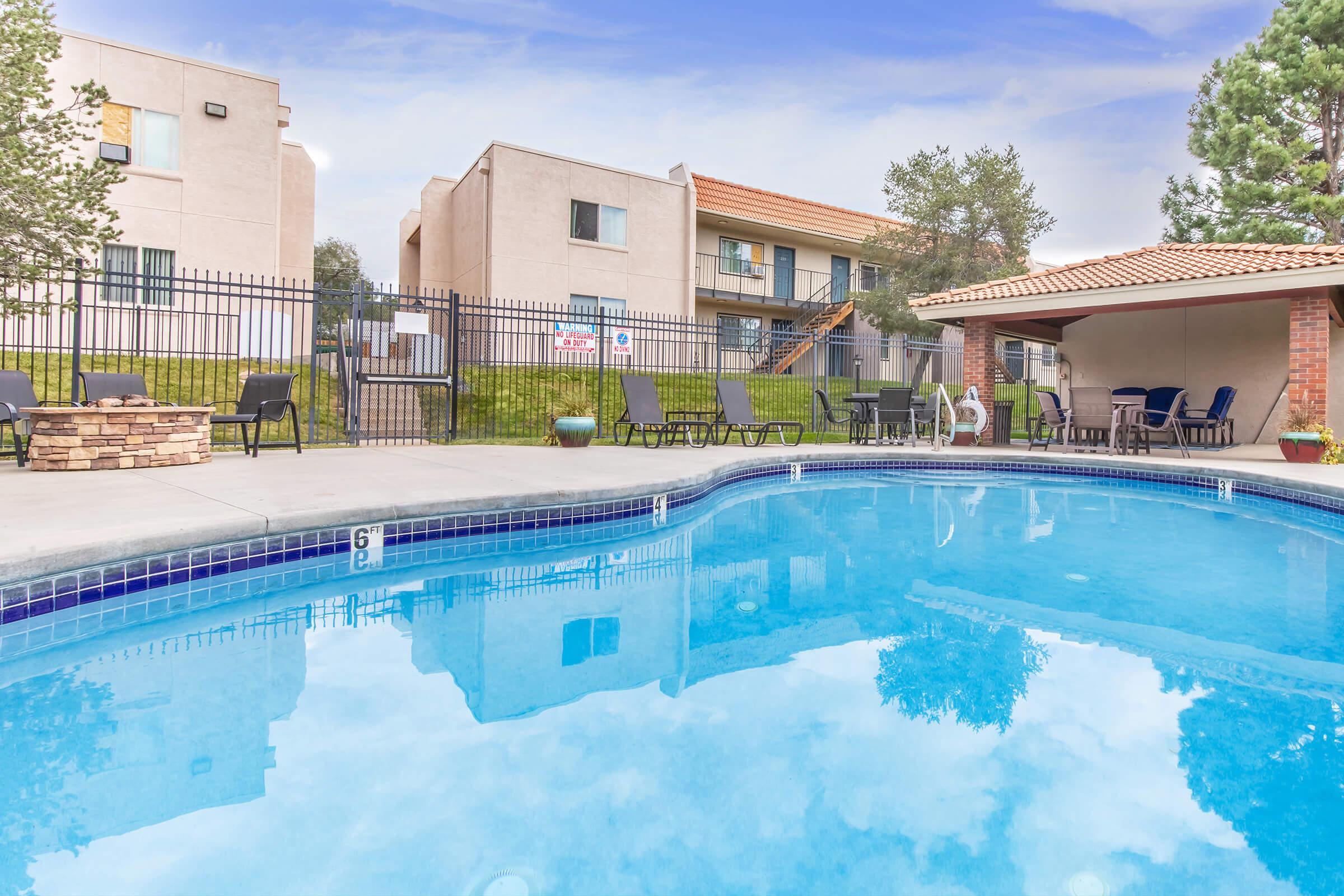 A clear, blue swimming pool is surrounded by a fence, with lounge chairs and tables arranged nearby. In the background, there are several apartment buildings with a grassy area. Trees are visible around the pool area, creating a relaxing outdoor environment.