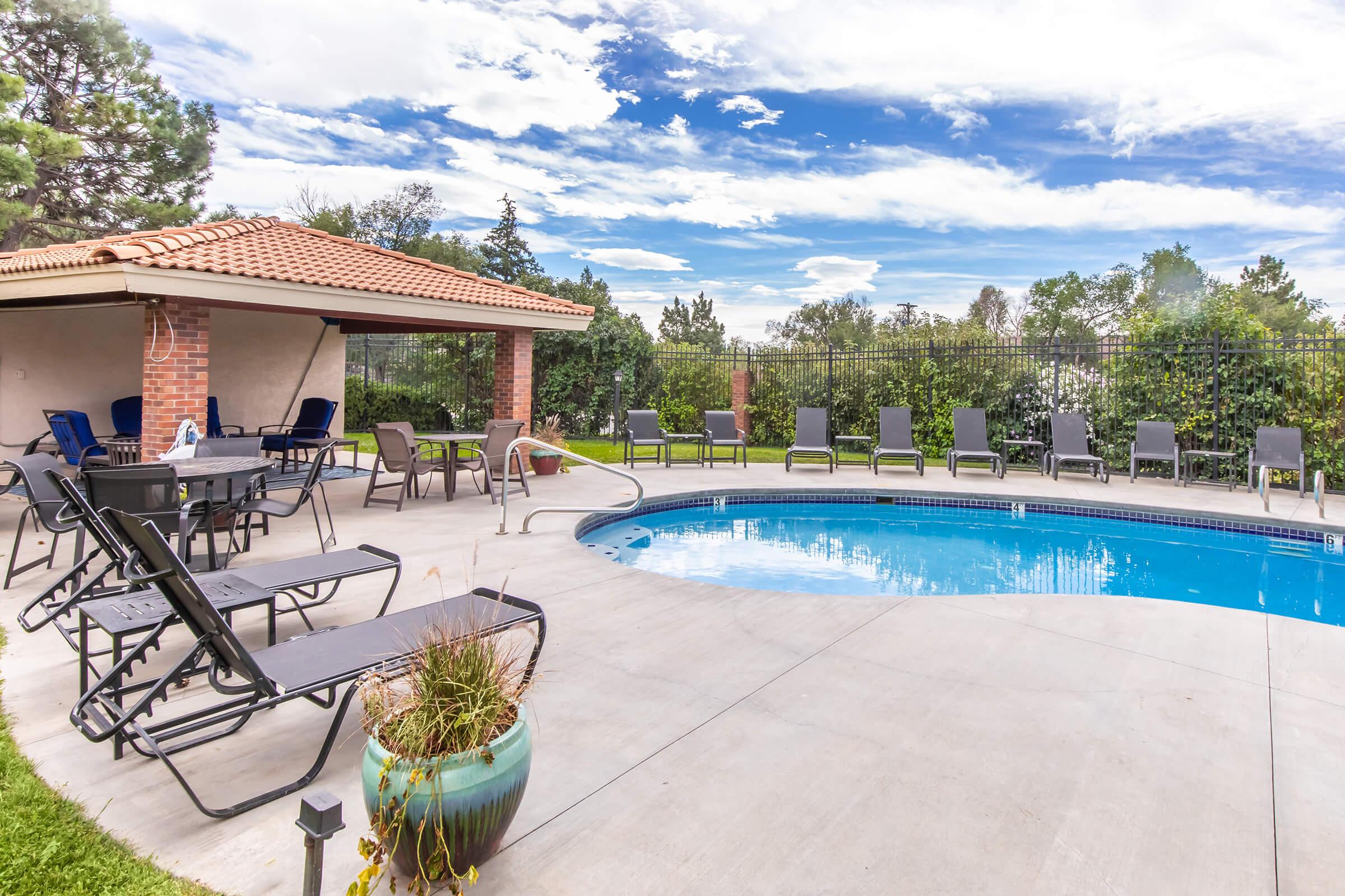 A swimming pool with clear blue water surrounded by lounge chairs under a gazebo. Lush greenery and trees are visible in the background, with a bright blue sky dotted with clouds. The area appears inviting and well-maintained, perfect for relaxation.