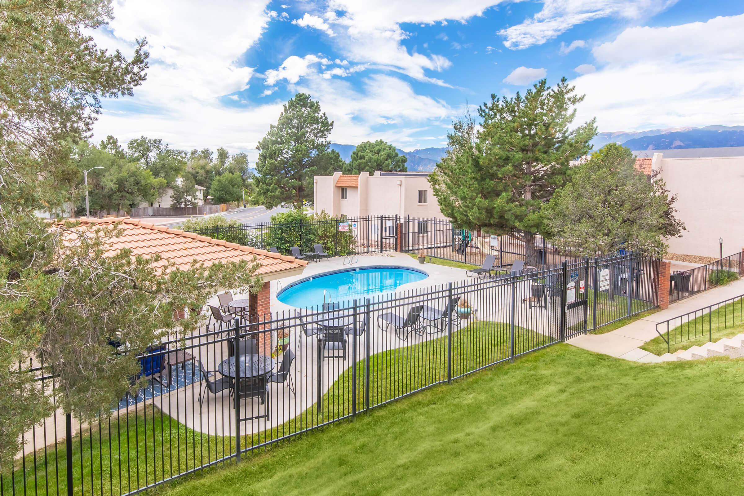 A view of a swimming pool surrounded by a black fence, with lounge chairs arranged nearby. There is a shaded area with a roof in the background, and lush green grass in the foreground. Trees provide some natural scenery, and a clear blue sky with scattered clouds can be seen overhead.