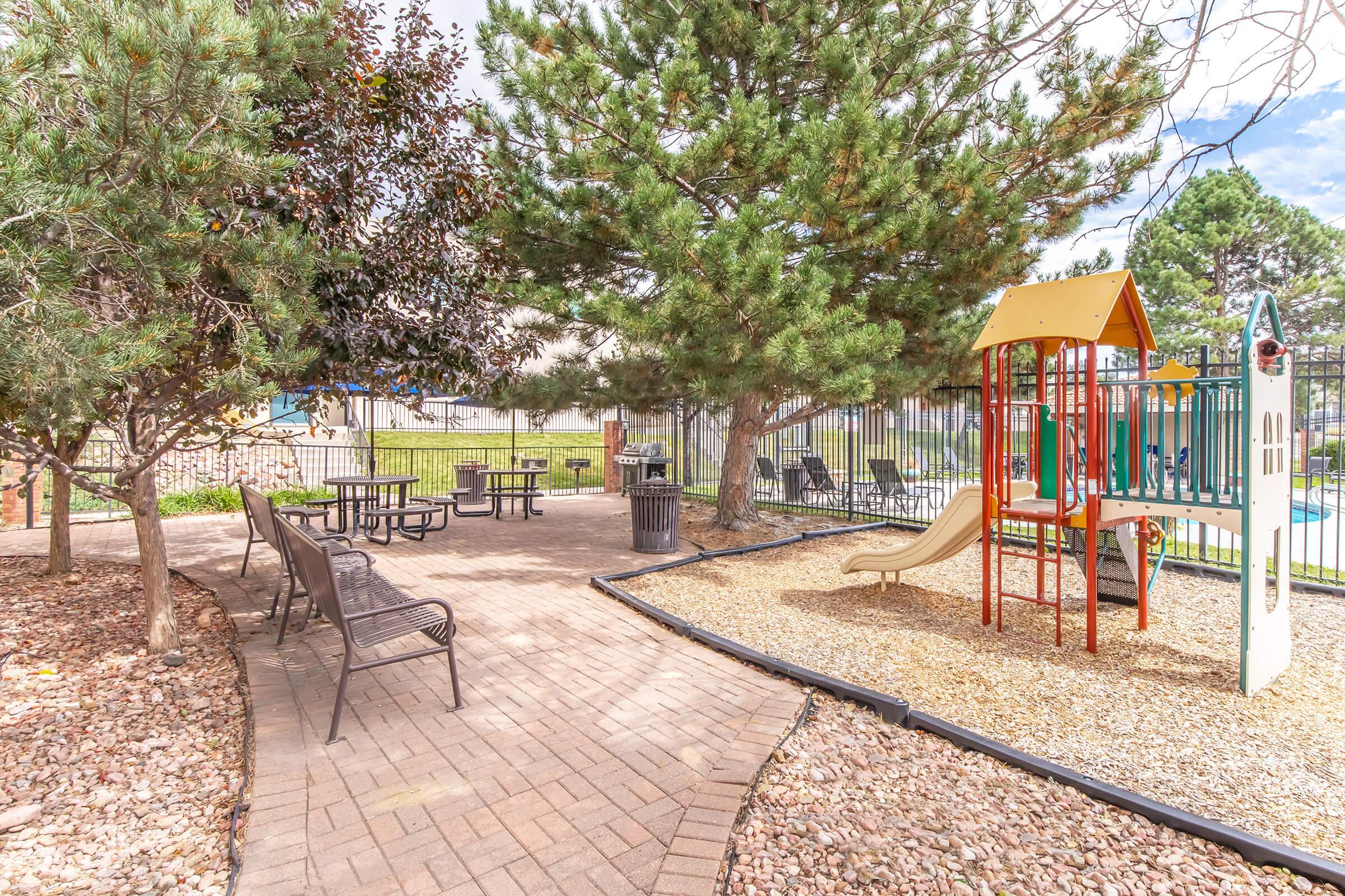A playground area featuring a colorful slide and climbing structure, surrounded by trees and benches. The pathway is made of pavers, leading to a picnic table area. In the background, there are more trees and a fence, with a glimpse of a pool area. The scene is bright and inviting, suitable for family activities.