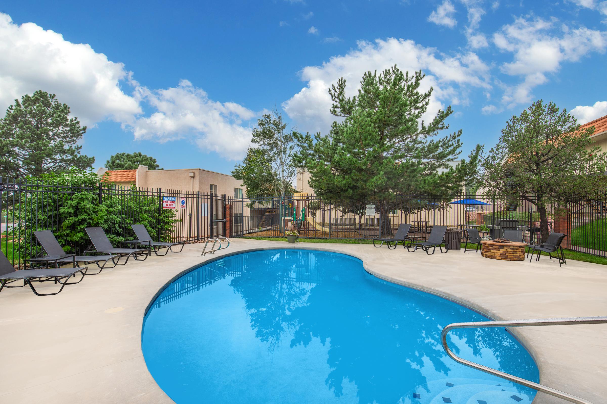 A serene outdoor swimming pool surrounded by lounge chairs, with lush green trees and a clear blue sky in the background. The pool features a gentle curve and a railing for entry, while a fenced area provides privacy. Nearby, there's a fire pit set in a landscaped space.