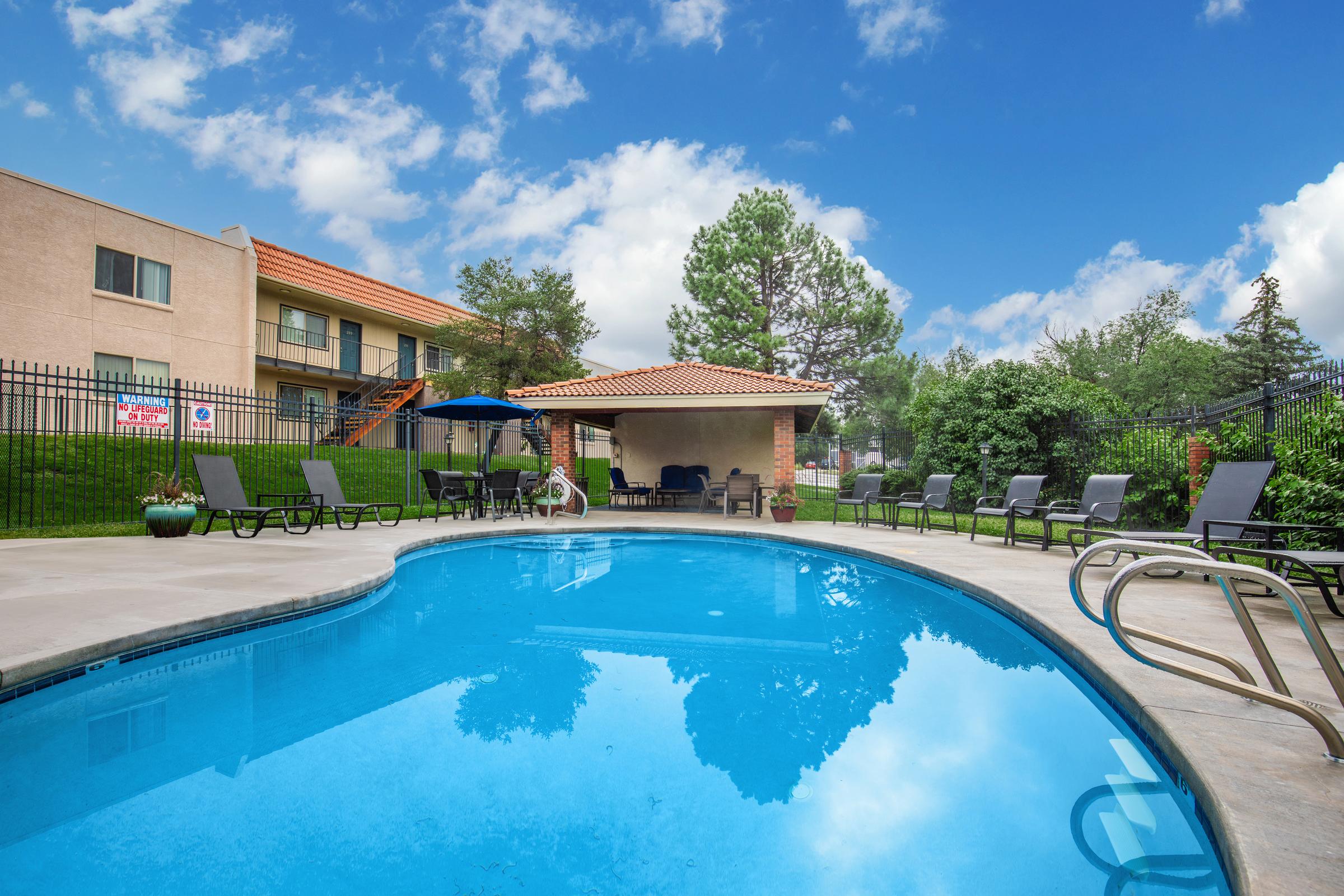 A clear blue swimming pool surrounded by lounge chairs, with lush greenery and a fence in the background. There’s a shaded area with tables and umbrellas, and a building with a terracotta roof is visible. The sky is bright with scattered clouds, creating a relaxing outdoor atmosphere.