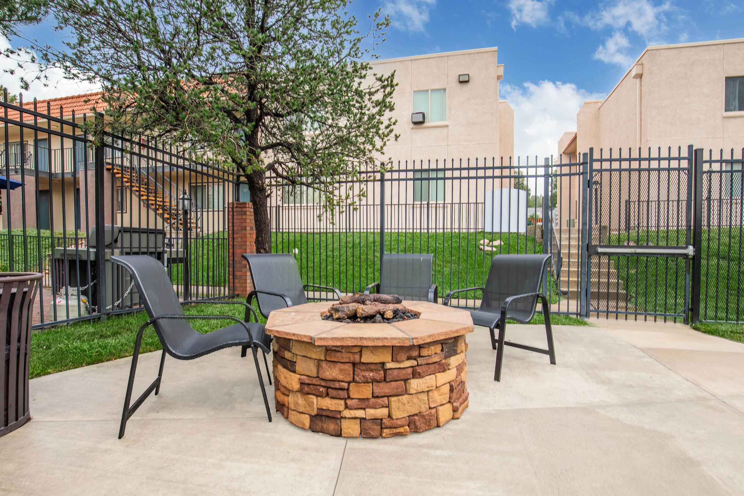 A cozy outdoor fire pit area with a circular stone seating arrangement, surrounded by black fencing and green grass. In the background, there are two beige apartment buildings and a tree providing shade. The sky is partially cloudy, creating a welcoming atmosphere for gatherings.