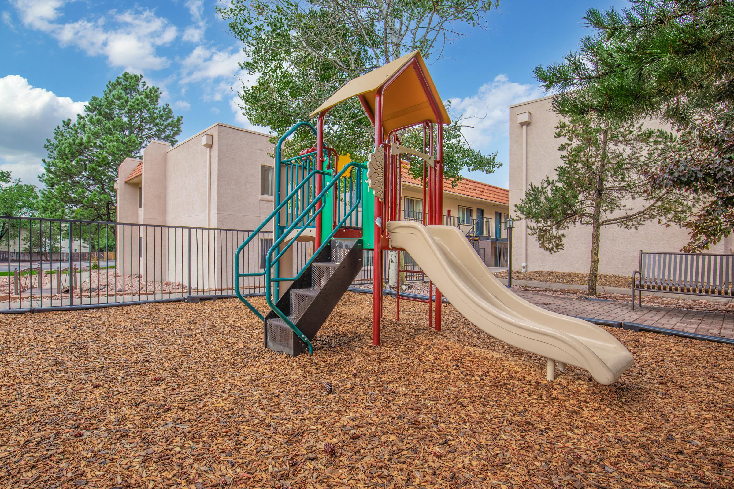 Playground structure featuring a yellow slide and climbing stairs, situated on a bed of wood chips. In the background, there are several buildings and trees under a partly cloudy blue sky. The area is enclosed by a fence, creating a safe play environment for children.