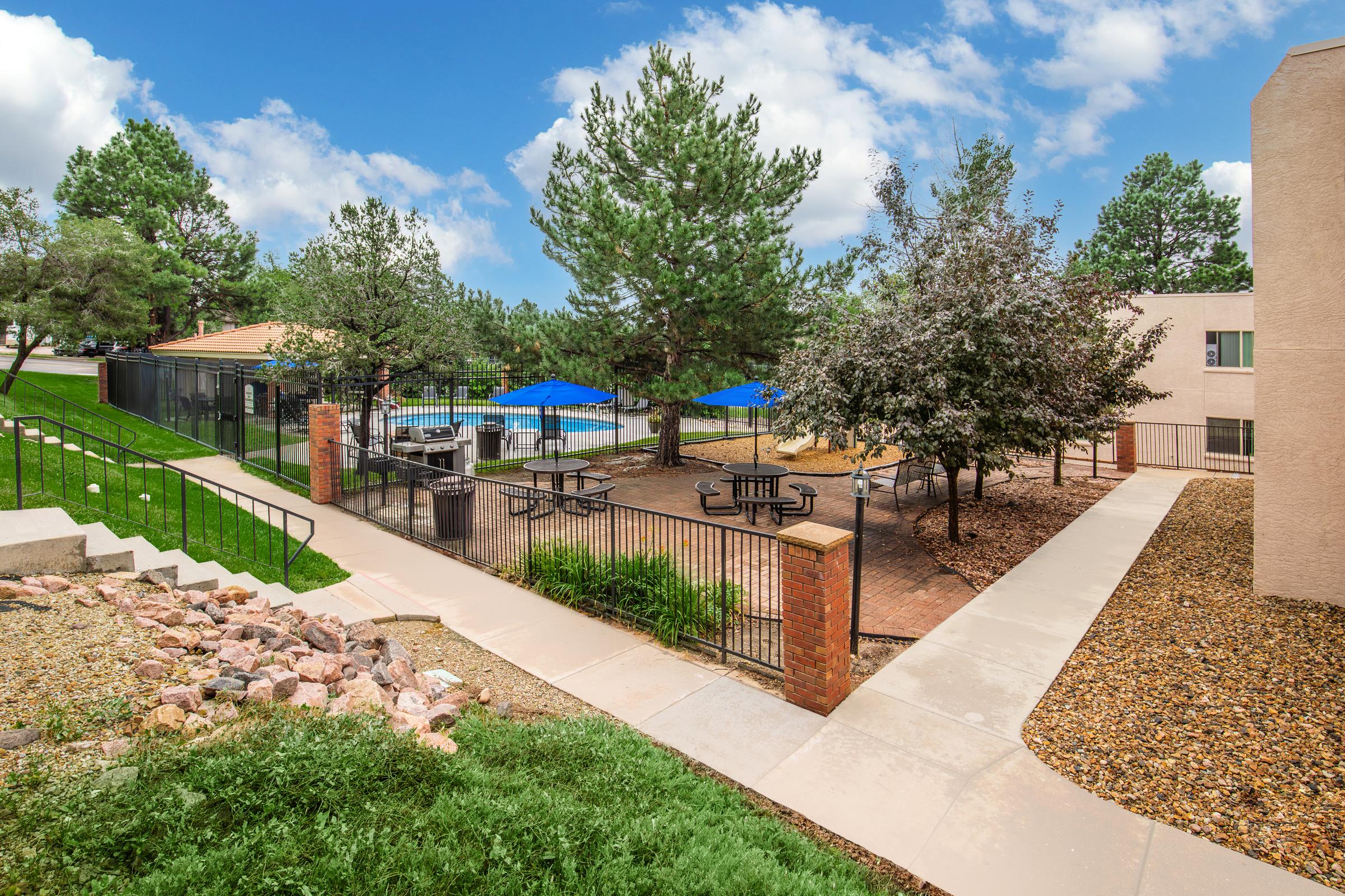 View of a landscaped outdoor area featuring a swimming pool surrounded by lounge seating and blue umbrellas. There are picnic tables and benches on a brick patio, with trees and grassy areas nearby. Walking paths made of concrete and gravel lead through the space, showcasing a serene and inviting atmosphere.