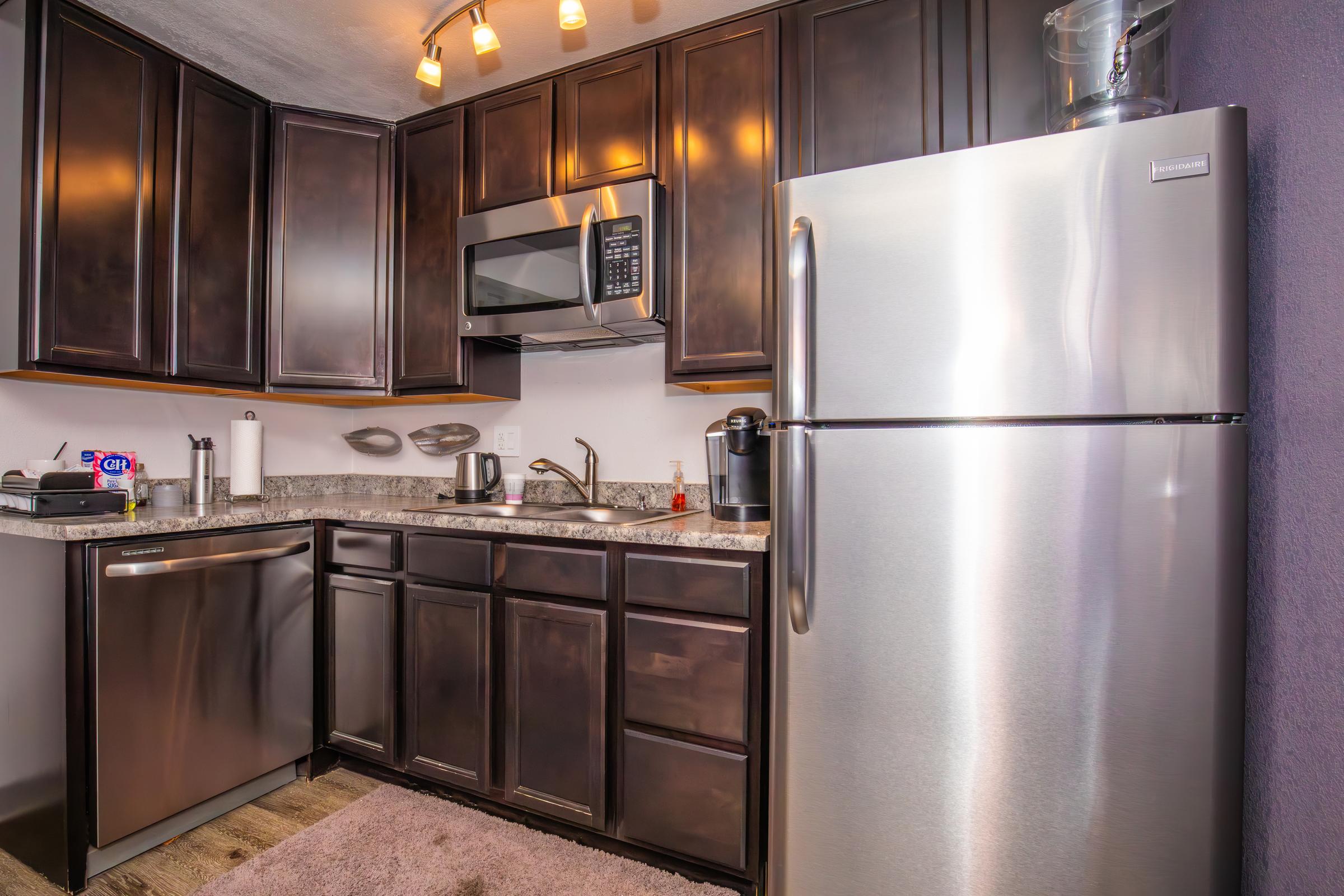 Modern kitchen featuring dark wooden cabinets, stainless steel appliances including a refrigerator and microwave, a granite countertop, and a small coffee maker. The space is well-lit with track lighting and includes a plush rug on the floor.