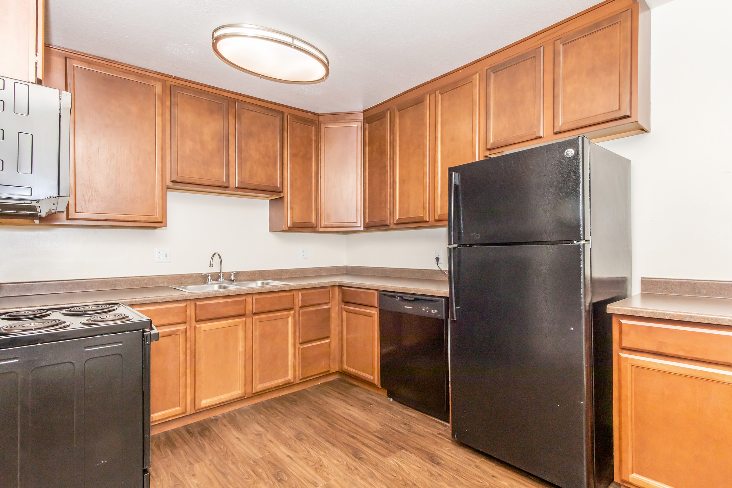 A modern kitchen featuring wooden cabinets, a black refrigerator, a dishwasher, and a stove. The countertop is a neutral color, and the floor is a wood-like laminate. The ceiling has a round light fixture, providing a bright and functional cooking space.