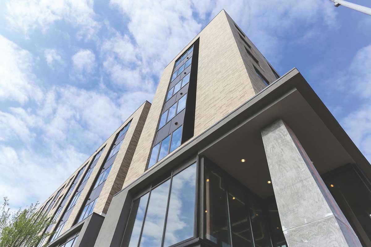 A modern building with a mix of brick and glass facades, viewed from a low angle. The structure features large windows reflecting the sky, and a cloudy blue backdrop. The scene includes greenery at the base, suggesting a contemporary urban environment.