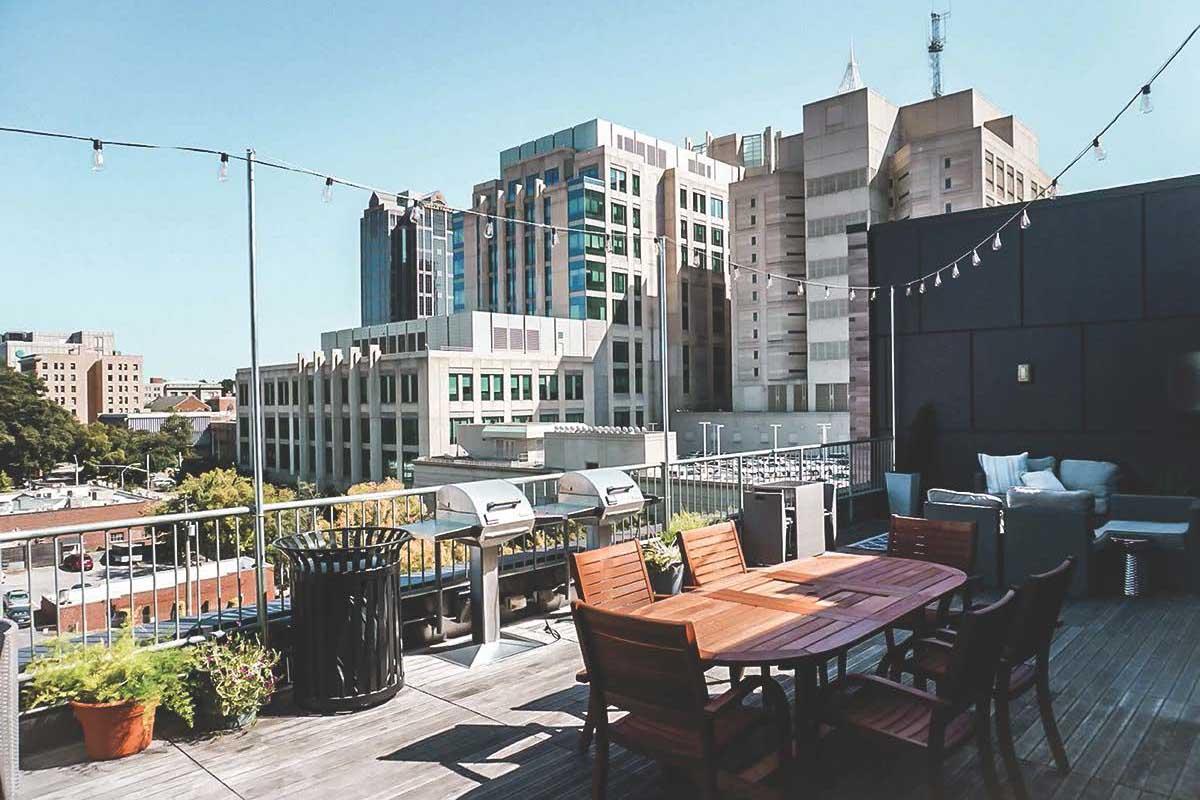 A rooftop terrace featuring a wooden dining table with chairs, barbecue grills, potted plants, and a view of city buildings. String lights are hanging above, and the sky is clear blue, creating a relaxed outdoor atmosphere.