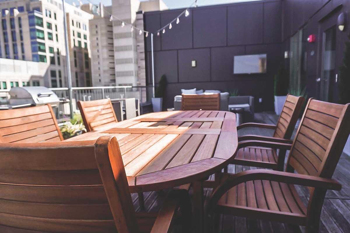 A wooden outdoor dining table surrounded by matching chairs on a rooftop terrace. In the background, modern buildings are visible, and there are decorative string lights overhead. A lounge area with a TV is also partially visible, creating a relaxed atmosphere.