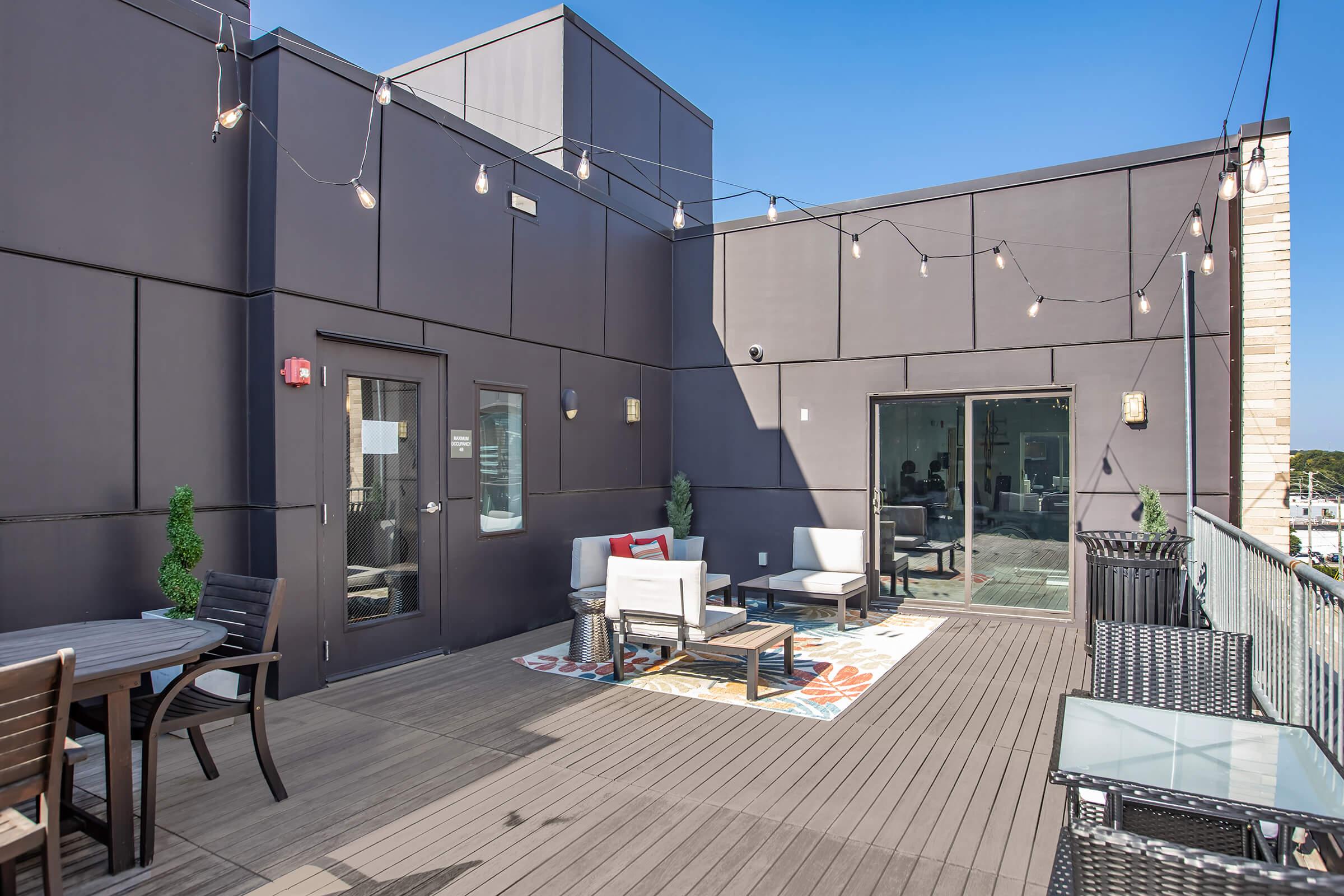 A spacious rooftop terrace featuring modern outdoor furniture, including a seating area with white chairs and tables, surrounded by decorative plants. The space is illuminated with string lights, offering a clear blue sky in the background, creating a relaxed and inviting atmosphere.