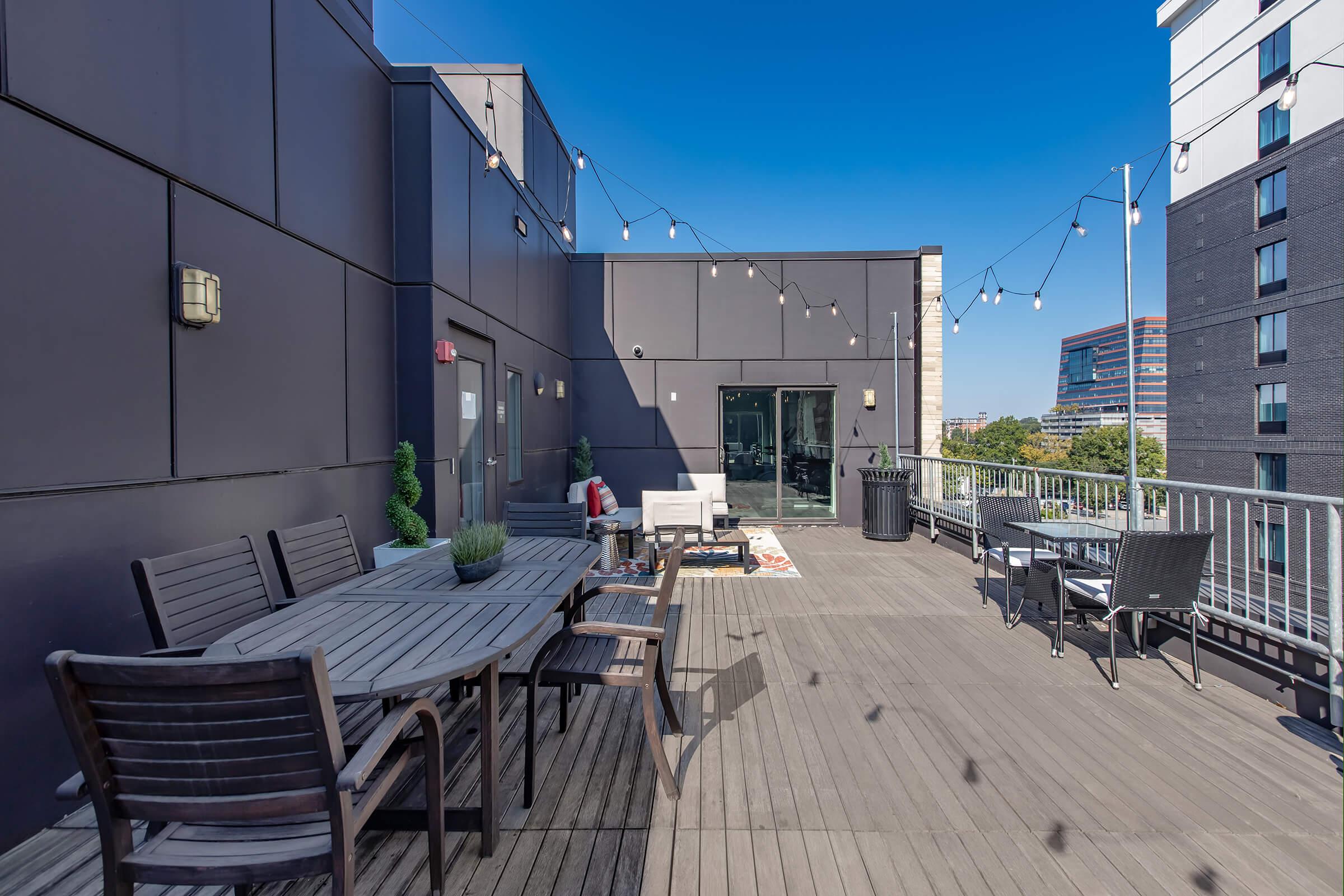 A rooftop terrace featuring wooden flooring, a set of dining tables and chairs, and lounge seating. The space is adorned with string lights, surrounded by modern buildings, and has a clear blue sky in the background. This area offers a stylish outdoor setting for relaxation and social gatherings.
