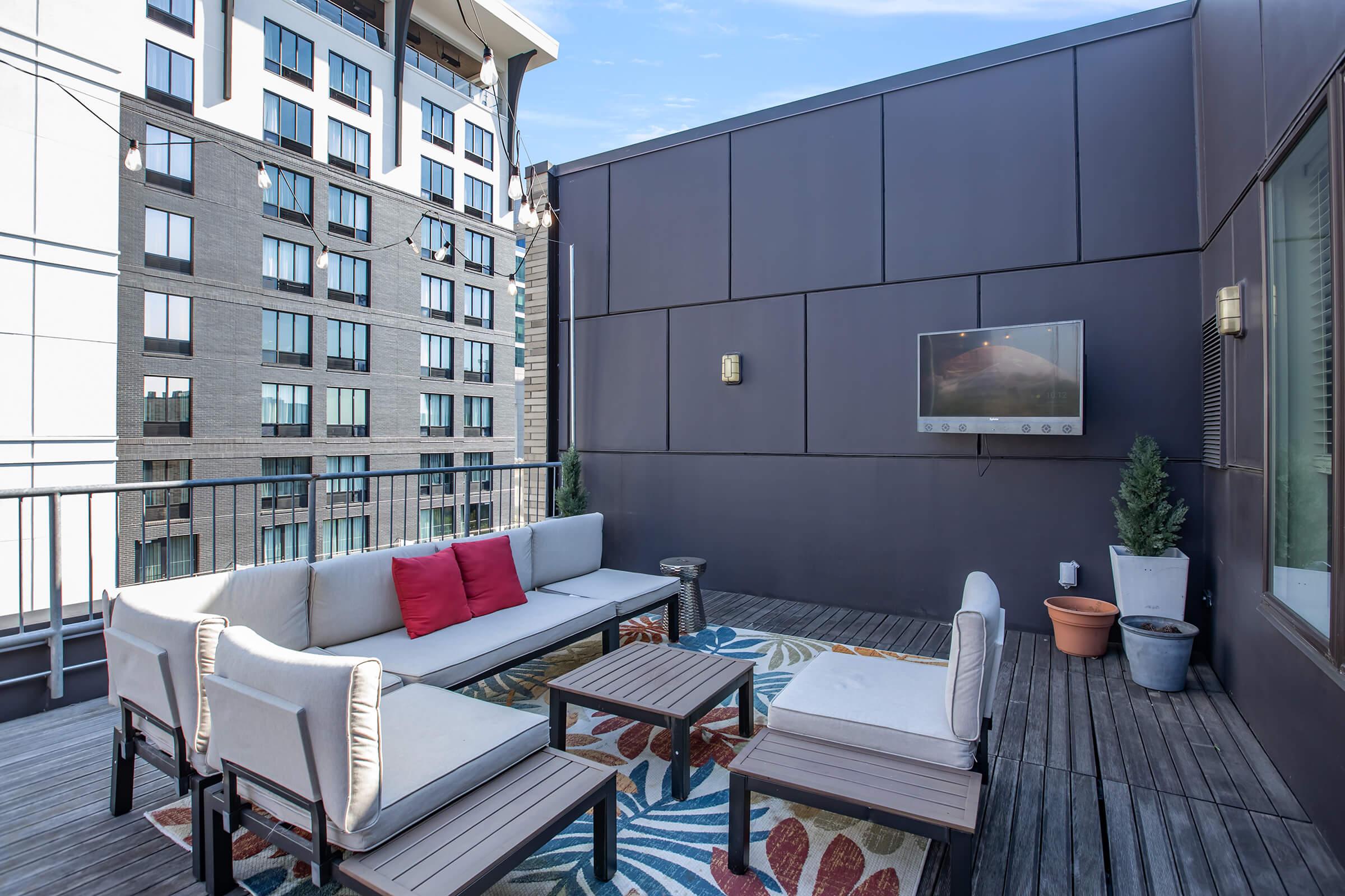 A modern rooftop terrace featuring a comfortable seating area with a sectional sofa and two chairs, a coffee table, and a flat-screen TV mounted on the wall. The space has a colorful area rug and potted greenery, with a backdrop of a multi-story apartment building and blue sky.