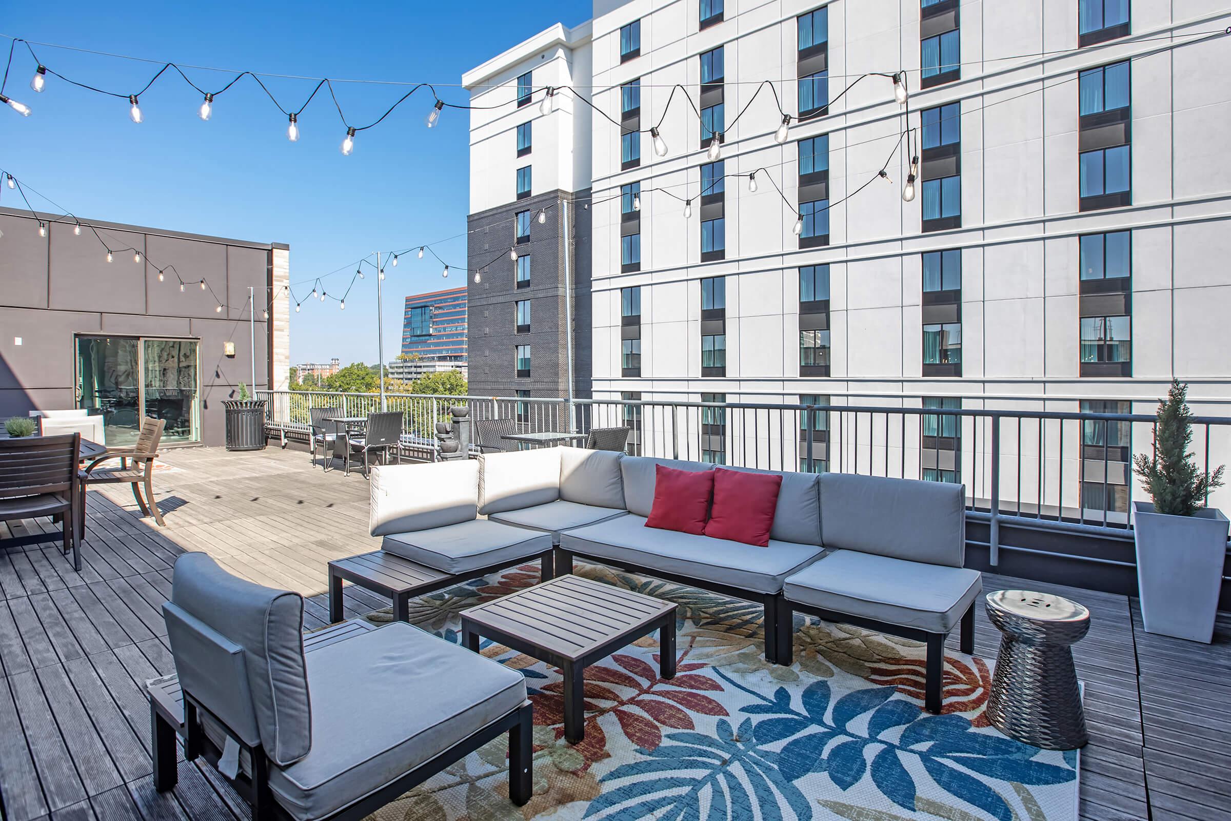 Rooftop lounge area featuring outdoor seating with gray cushions and a red decorative pillow, a wooden coffee table, and colorful area rug. String lights are hung above, and modern buildings are visible in the background under a clear blue sky.