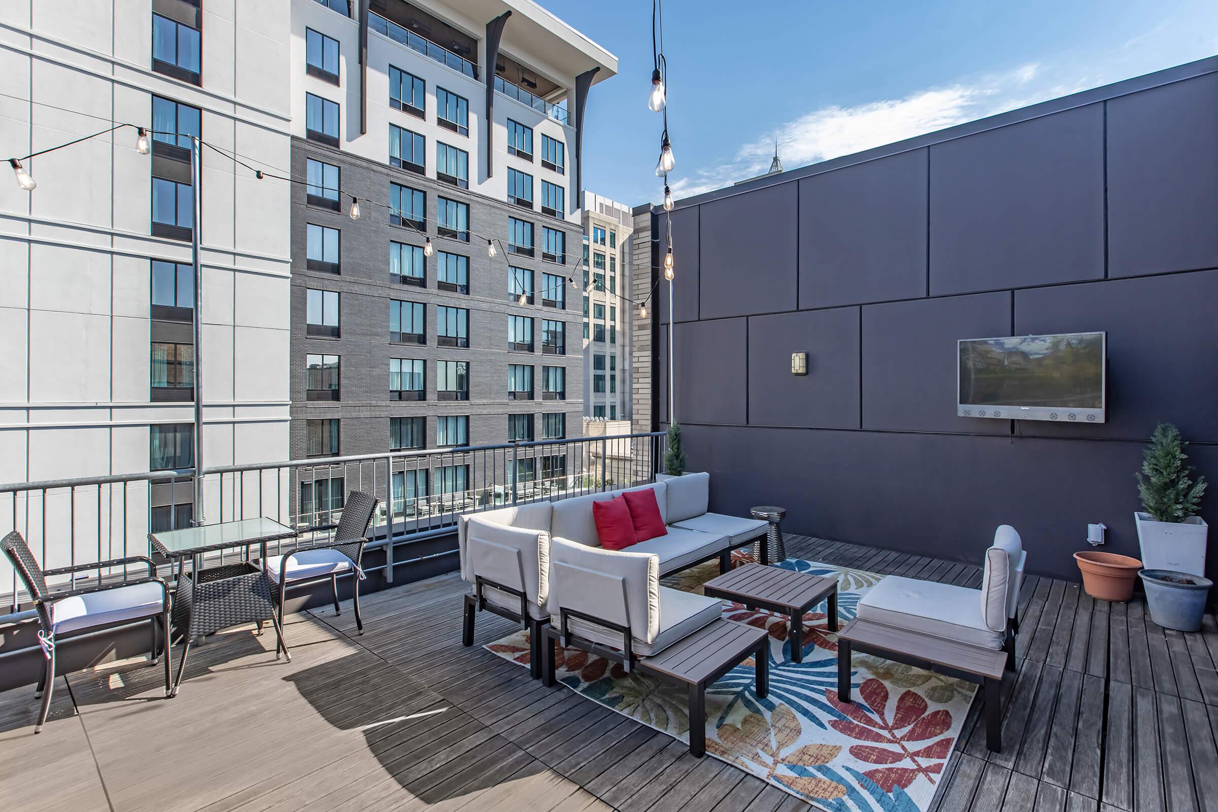 A stylish rooftop terrace featuring modern outdoor furniture, including a sofa with colorful cushions, a coffee table, and a dining area. Decorative string lights hang above, while a flat-screen TV is mounted on the wall. The backdrop consists of contemporary apartment buildings and a clear blue sky.