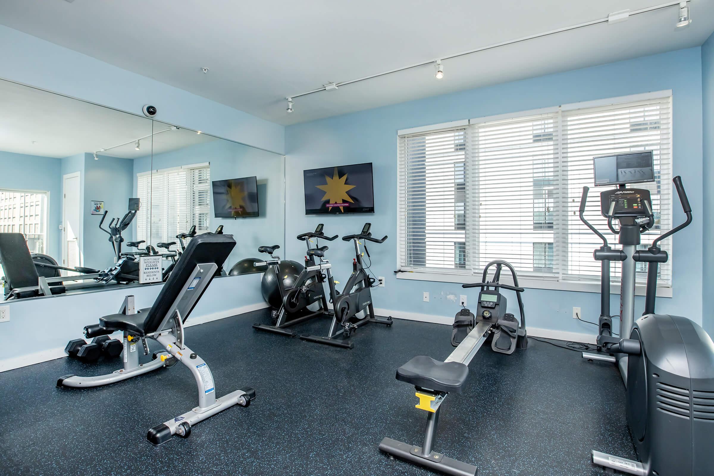 A well-lit gym interior featuring exercise equipment, including a stationary bike, a rowing machine, and a weight bench. The room has large windows with blinds, mirrors on the wall, and a television mounted above the equipment, all set against a light blue wall.