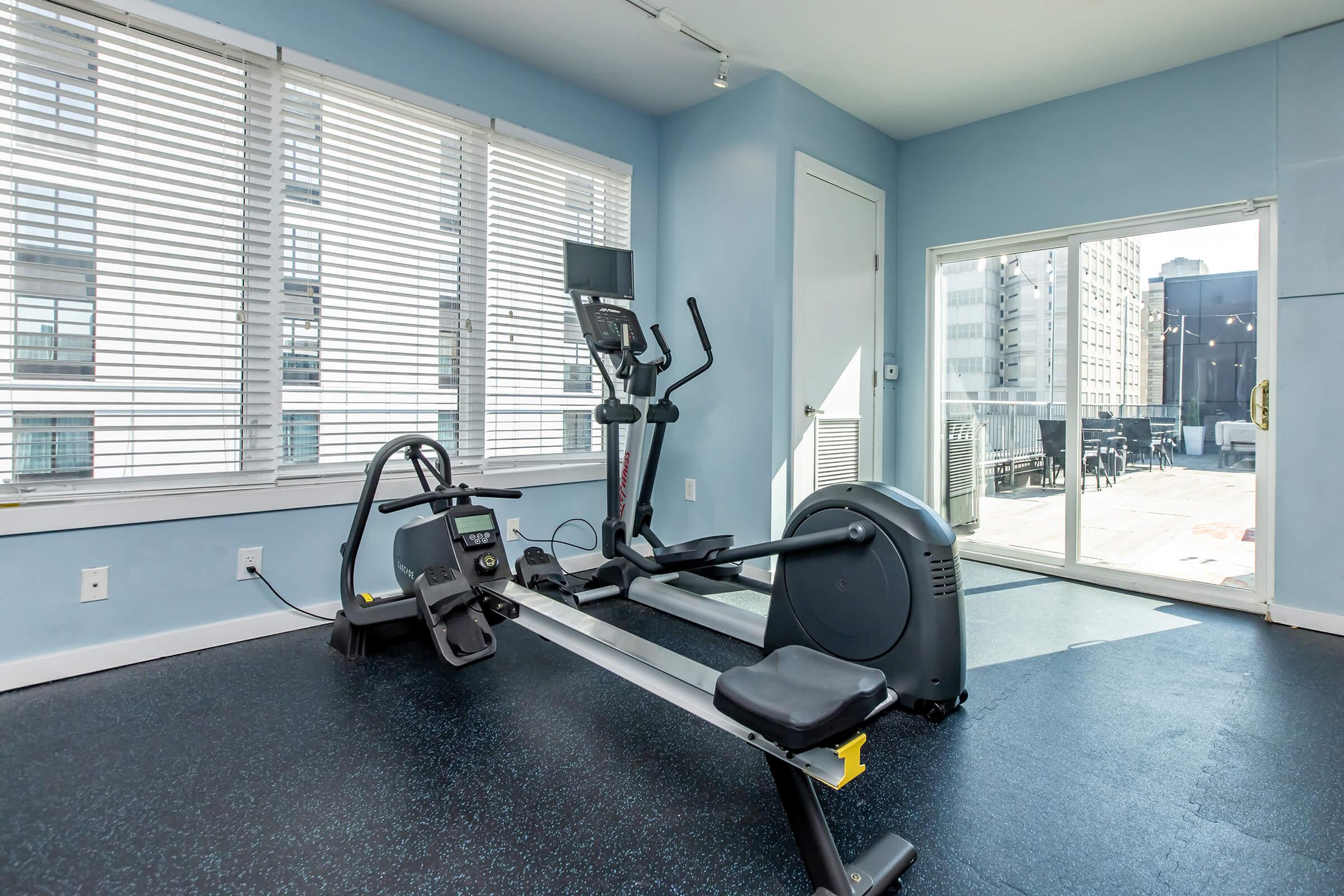 A well-lit fitness room featuring an elliptical machine and a rowing machine. Large windows provide natural light, and there is a view of an outdoor area. The floor is covered with dark exercise mats, creating a comfortable workout space.