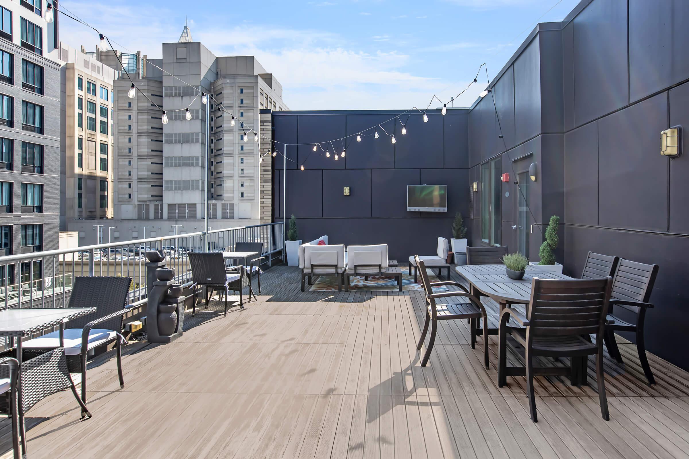 A rooftop terrace featuring wooden decking, outdoor furniture including tables and chairs, and decorative plants. String lights hang overhead, creating a cozy atmosphere. In the background, modern buildings can be seen against a clear sky, along with a wall-mounted television.