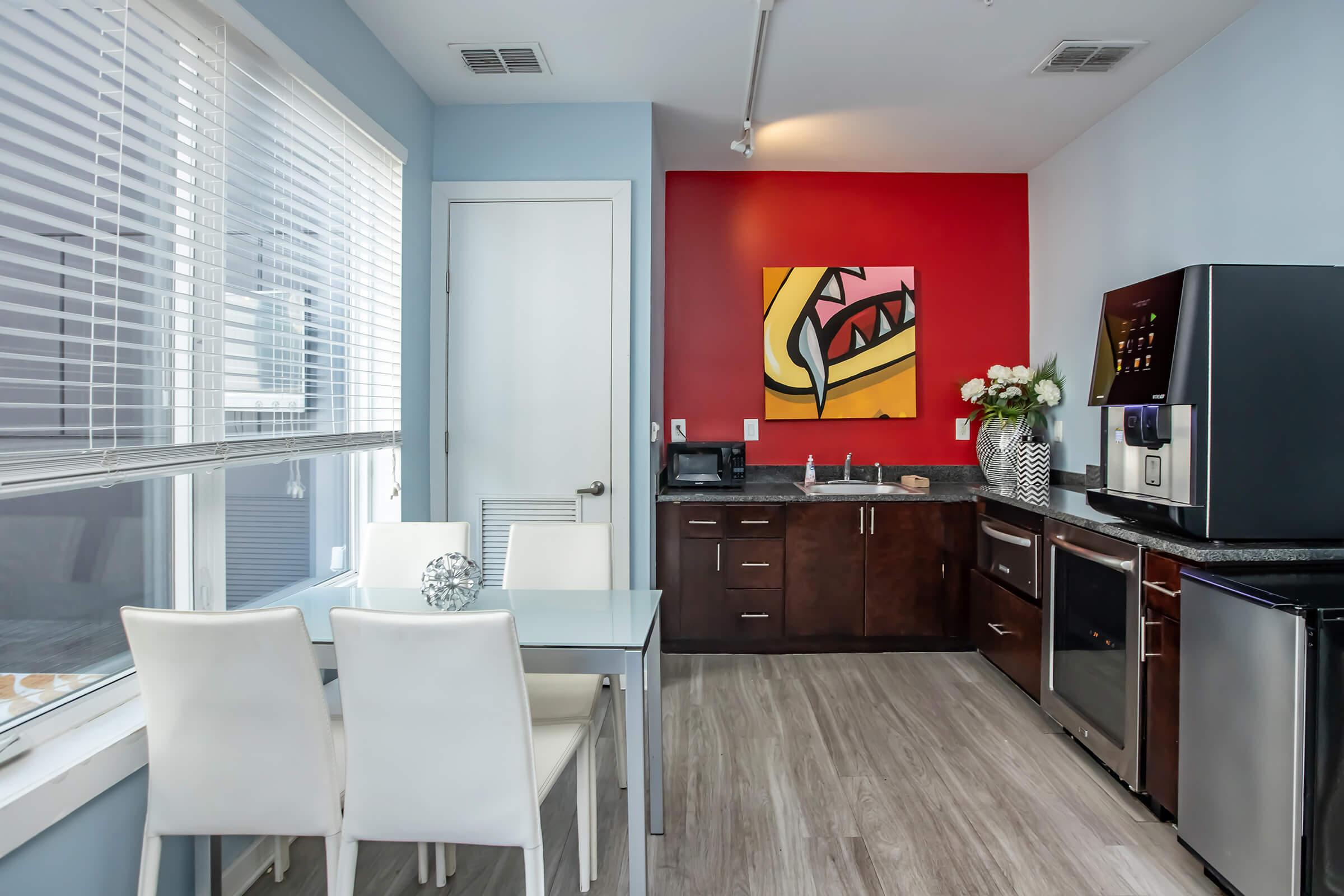 Modern kitchen with a sleek dark wood cabinet, stainless steel appliances, and a black refrigerator. A glass dining table with white chairs is set up near a window with blinds. A colorful artwork featuring a stylized shark is mounted on a bright red wall, adding a vibrant touch to the space.