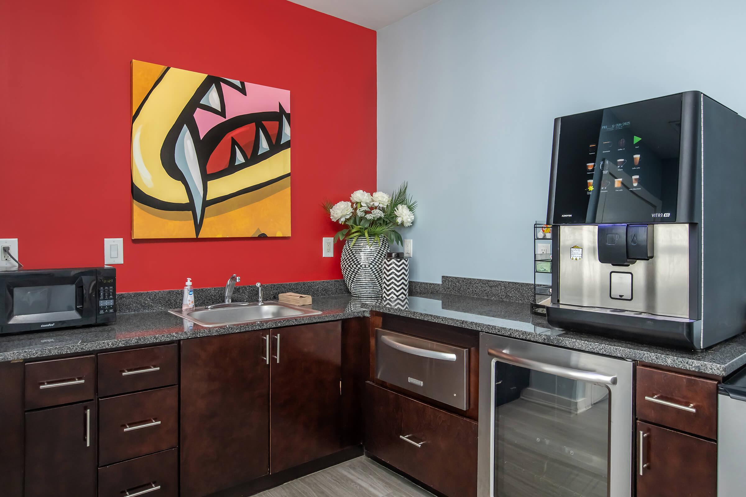 A modern kitchen showcasing dark wood cabinets and a sleek refrigerator with a water dispenser. There's a microwave and a small sink next to the refrigerator. A colorful artwork featuring a stylized creature with teeth hangs on the red wall above a countertop adorned with a vase of flowers.