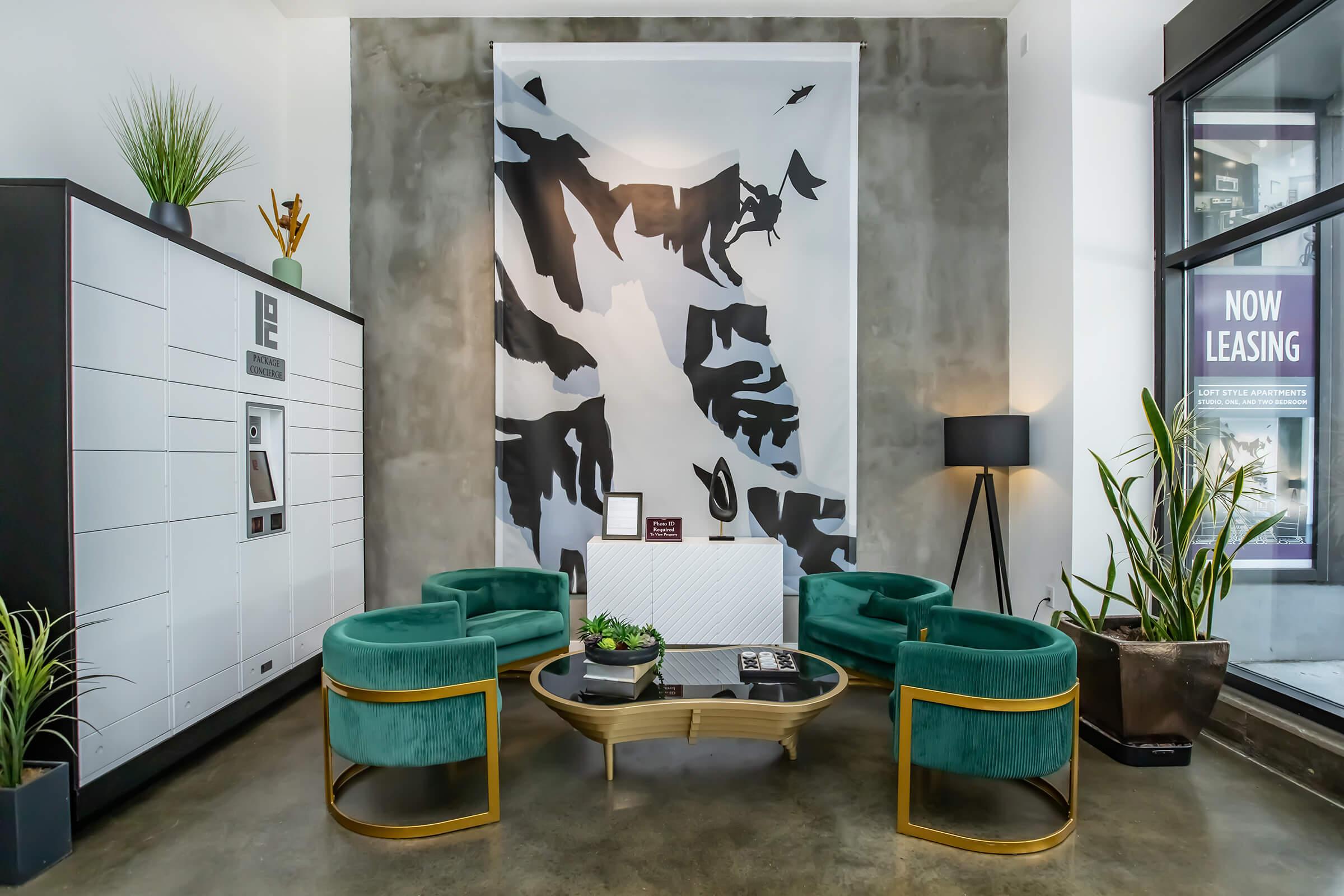 A modern interior space featuring four green velvet chairs arranged around a round coffee table. Behind the seating area, a large abstract black-and-white mural adorns the concrete wall. There's a decorative plant, a lamp, and a white storage unit visible, creating a stylish and inviting atmosphere.