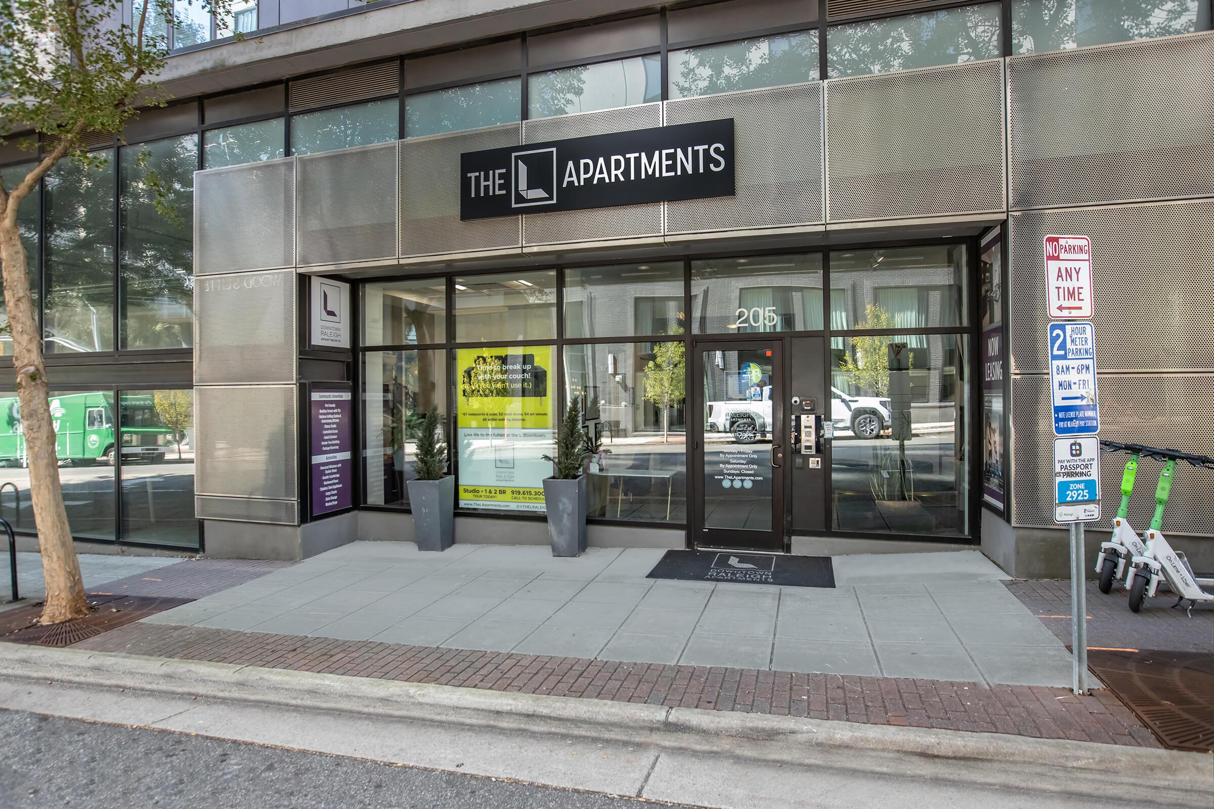 Entrance to "The L Apartments" building, featuring large glass doors and a modern facade. There are potted plants on either side of the entrance, and nearby, there are electric scooters parked. Visible street signs indicate parking regulations. The overall setting is urban and contemporary.