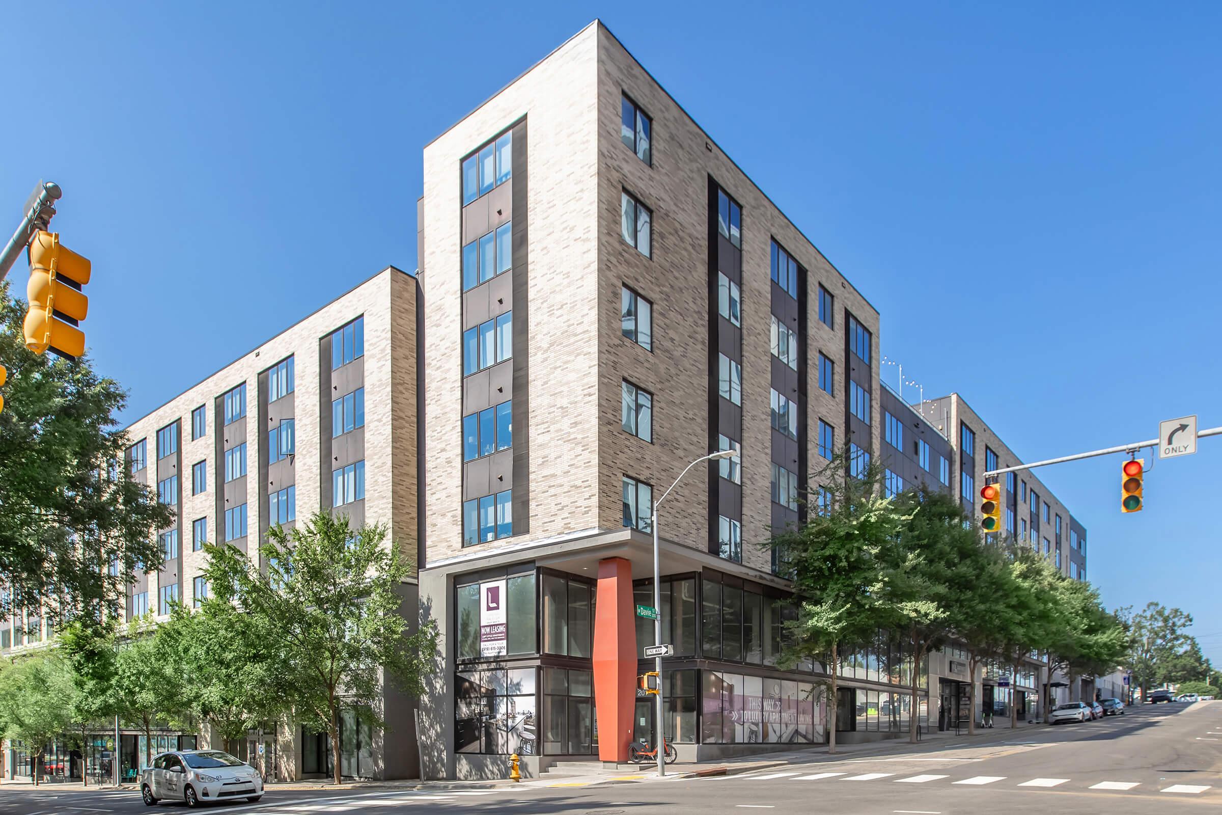 Modern multi-story building with a mix of brick and glass architecture. The structure features large windows, a prominent entrance, and is surrounded by trees. Traffic lights and a vehicle are visible on the street corner, indicating an urban setting. Clear blue sky in the background.