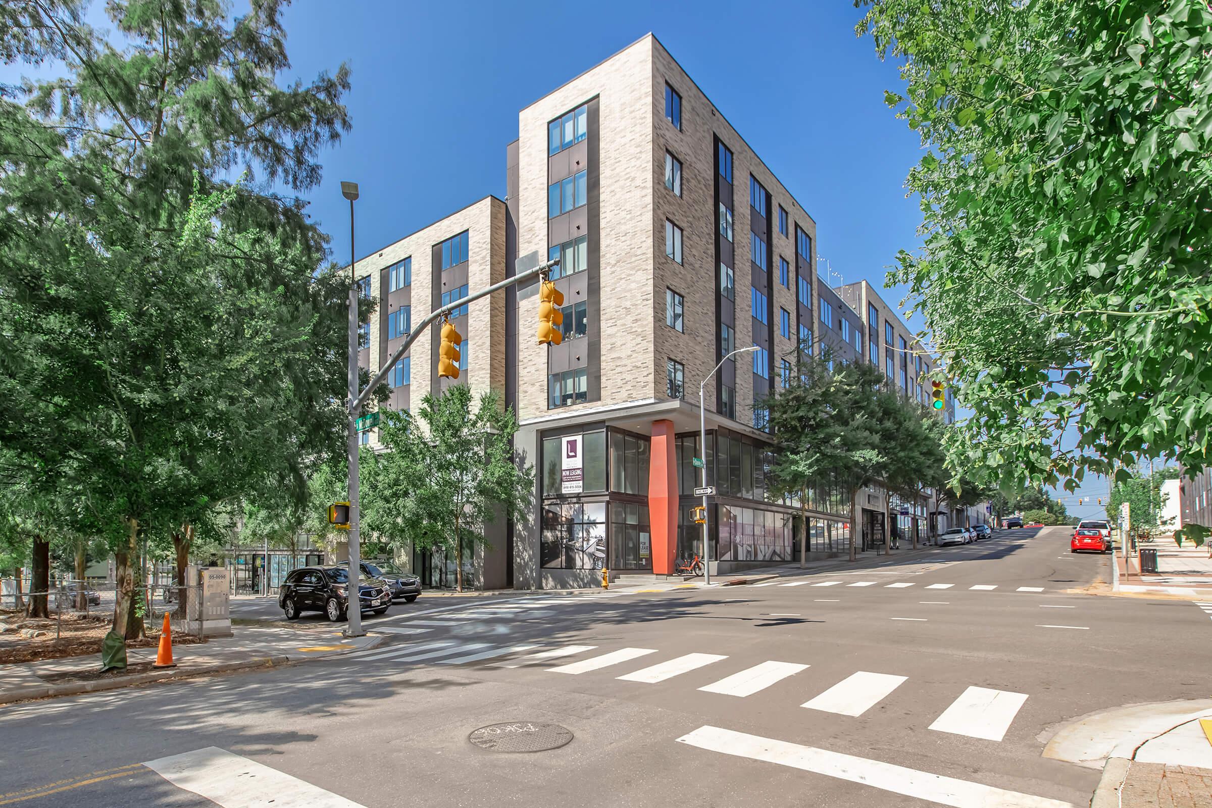 A modern, five-story apartment building at the corner of a city street, surrounded by trees. The building features large windows and a contemporary design, with a traffic light and crosswalk visible in the foreground. Clear blue sky provides a bright backdrop to the scene.