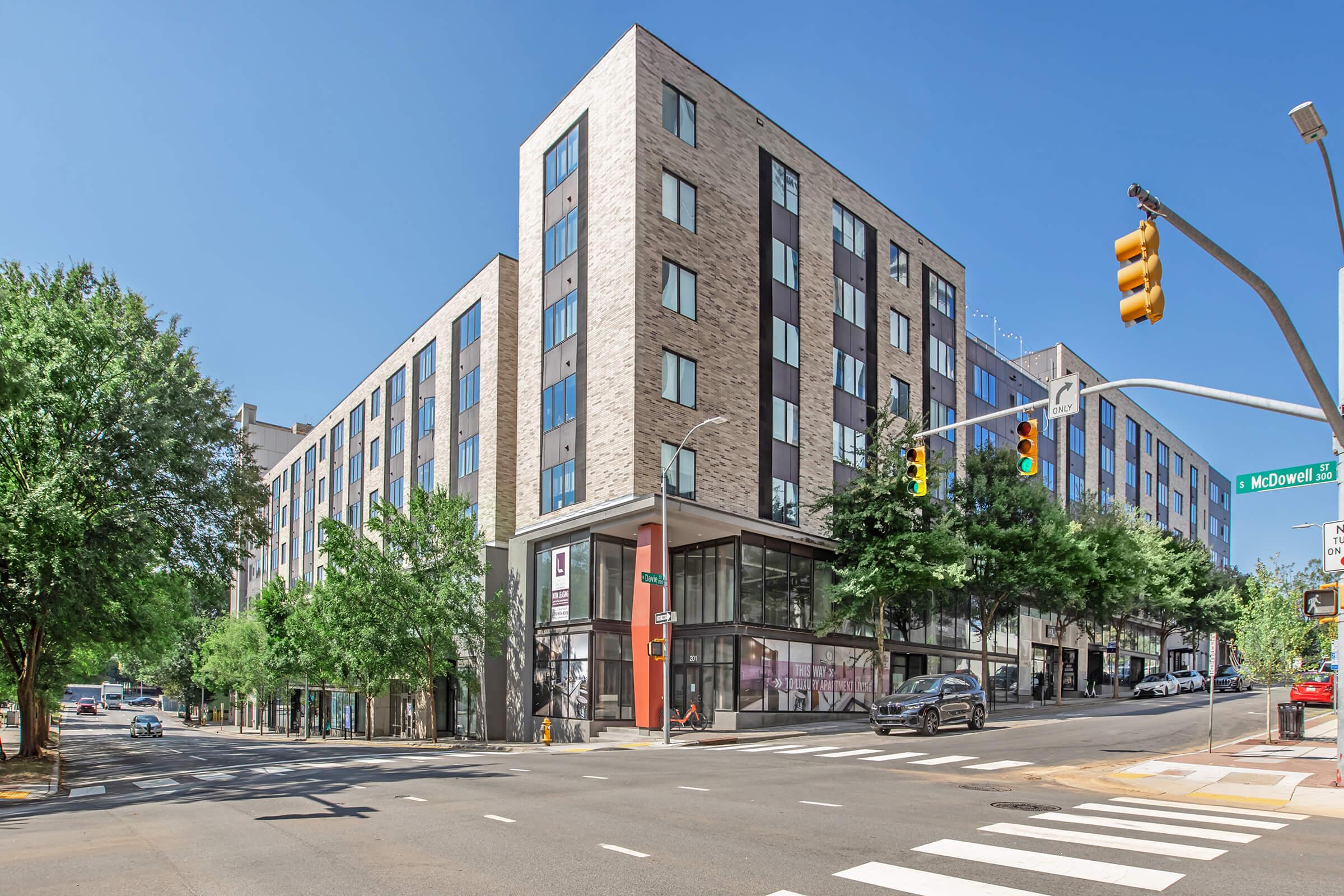 A modern multi-story building with a mix of brick and glass façade, located at a street intersection. It features large windows and tree-lined sidewalks, with vehicles and street signage visible in the foreground. The setting appears urban and well-maintained under clear blue skies.
