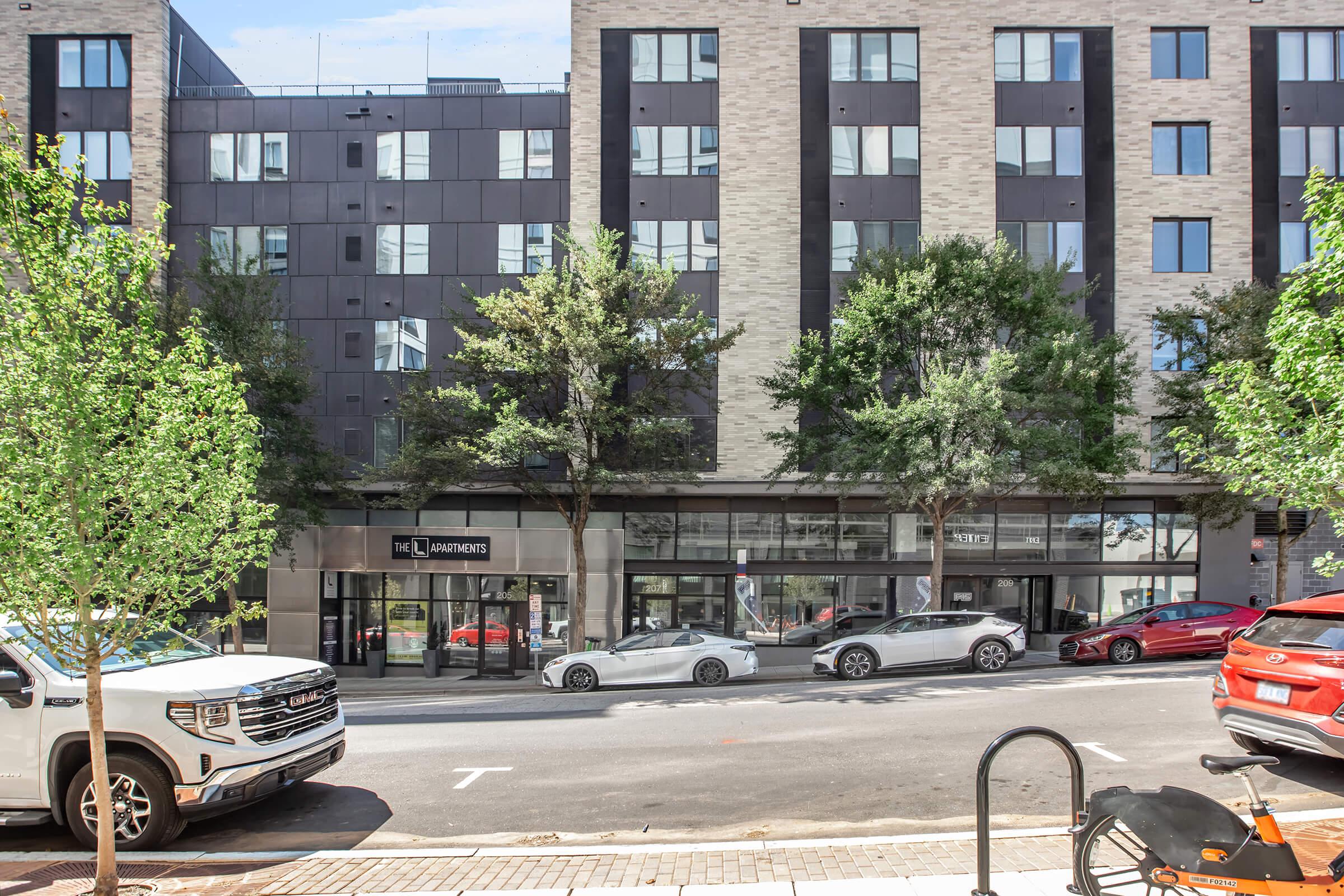 A street view of a modern apartment building with large windows and greenery in front. Several cars are parked along the street, including a white sports car and a red sedan. The building features a mix of brick and dark paneling, creating a contemporary aesthetic.