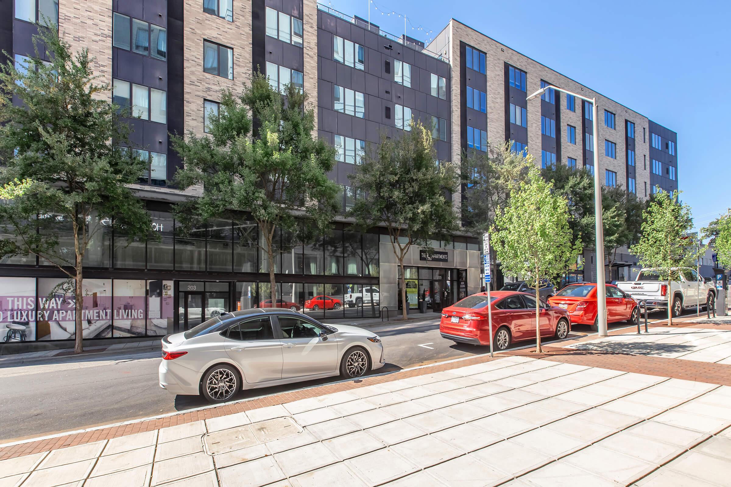 Urban scene featuring a row of parked cars, including a silver sedan and several red vehicles, along a wide sidewalk. In the background, modern multi-story buildings with large windows and a storefront. Clear blue sky above enhances the vibrant atmosphere of the street.
