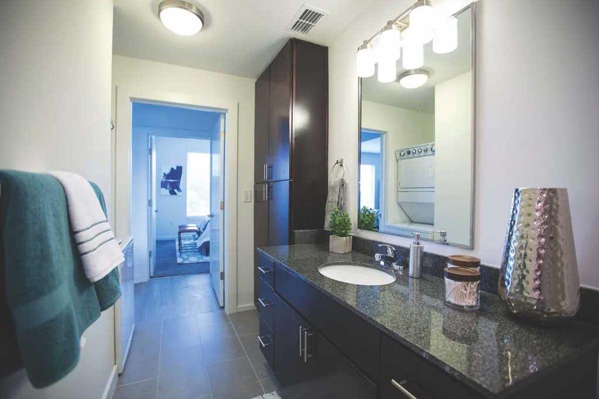 A modern bathroom featuring a dark wood vanity with a granite countertop and a round sink. Above the vanity is a large mirror and stylish light fixtures. A teal towel hangs on the side, and a decorative vase sits on the counter. The space is bright and clean, with a doorway leading to another room in the background.