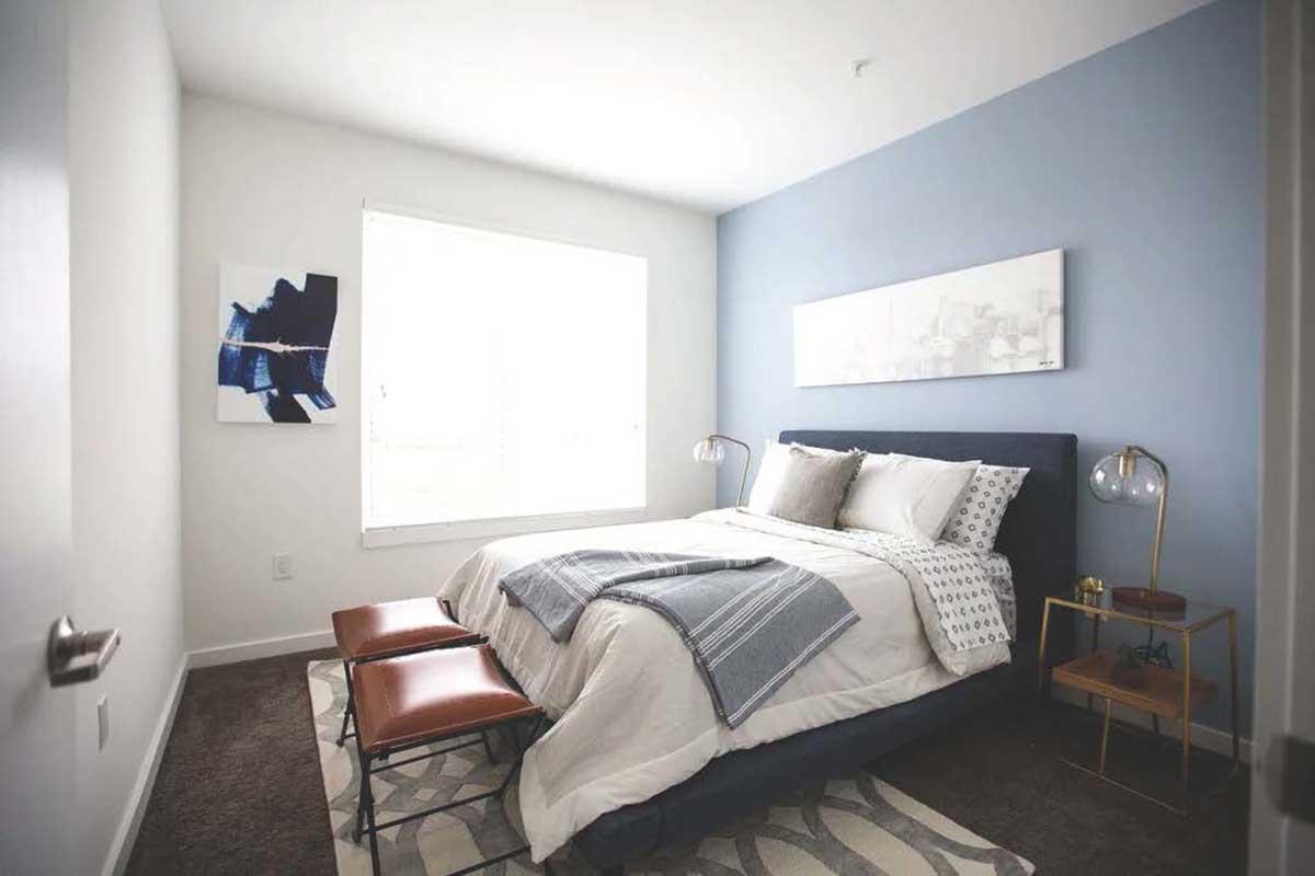 A cozy bedroom featuring a neatly made bed with white and gray bedding, a blue accent wall, two simple brown stools at the foot of the bed, and a compact side table with a lamp. Natural light streams in from a large window, enhancing the modern and inviting atmosphere of the space.