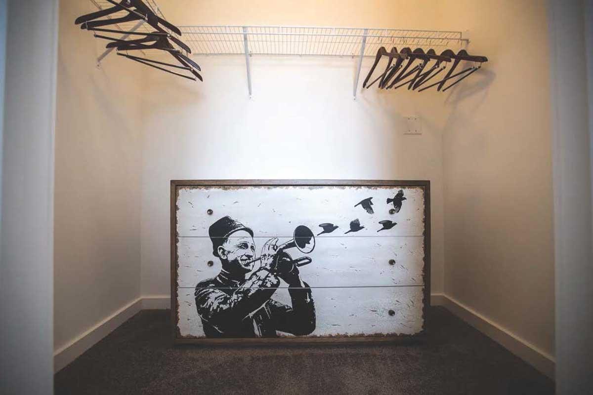 A minimalist closet featuring a white wall with a black and white mural of a man playing a trumpet, surrounded by silhouettes of flying birds. Above, there is a wire shelf with empty hangers, emphasizing the simplicity of the space. The floor is carpeted in a neutral tone.