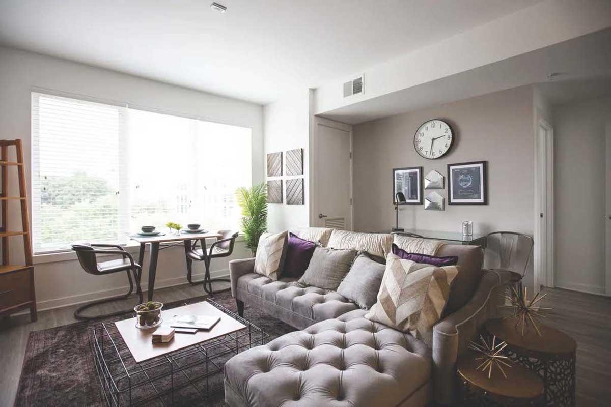 A modern living room featuring a gray tufted sofa with decorative pillows, a stylish coffee table with a magazine and plant, and a dining area with a round table and chairs. Large windows let in natural light, and minimalist decor includes wall art and a clock, creating a cozy and inviting atmosphere.
