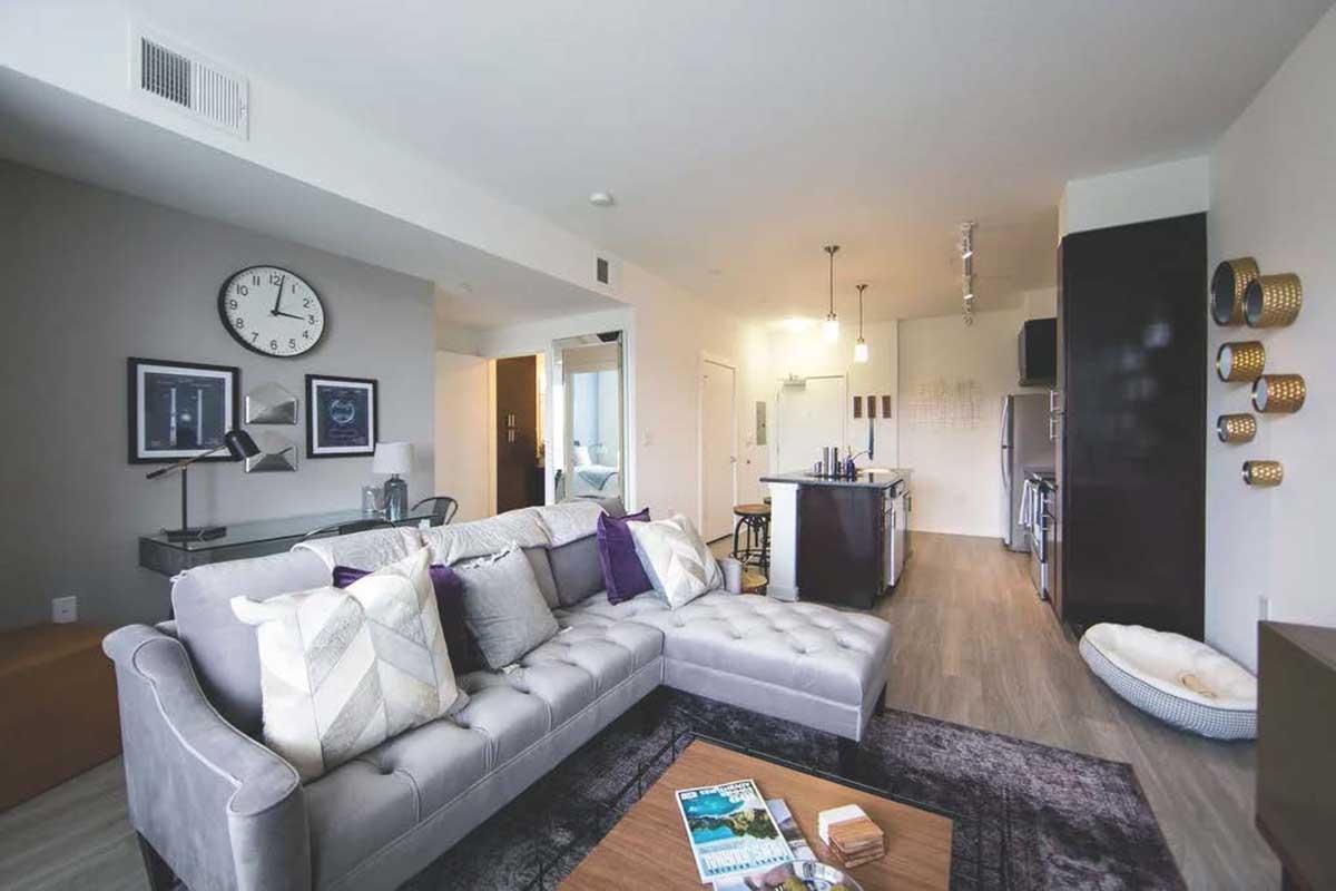 A modern living room featuring a gray sectional sofa with decorative pillows, a wooden coffee table, and a stylish rug. On the wall are framed artworks and a large clock. The open kitchen is visible in the background, with an island, bar stools, and contemporary appliances. Natural light filters in through the windows.