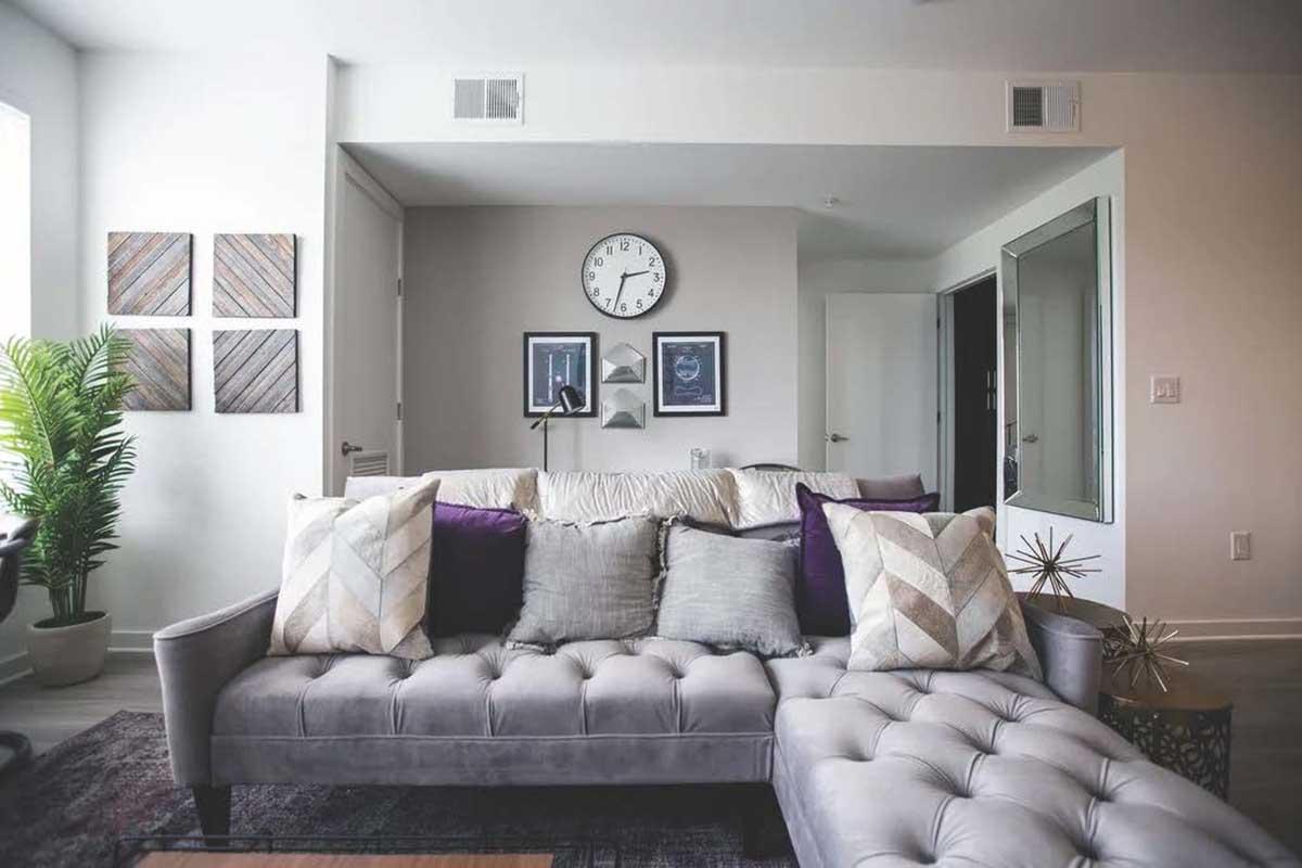 A modern living room featuring a gray tufted sofa adorned with decorative pillows. A circular wall clock is displayed above a pair of framed artworks. There's a contemporary table and a potted plant, along with a mirror reflecting part of the space. The room has a bright and airy atmosphere.