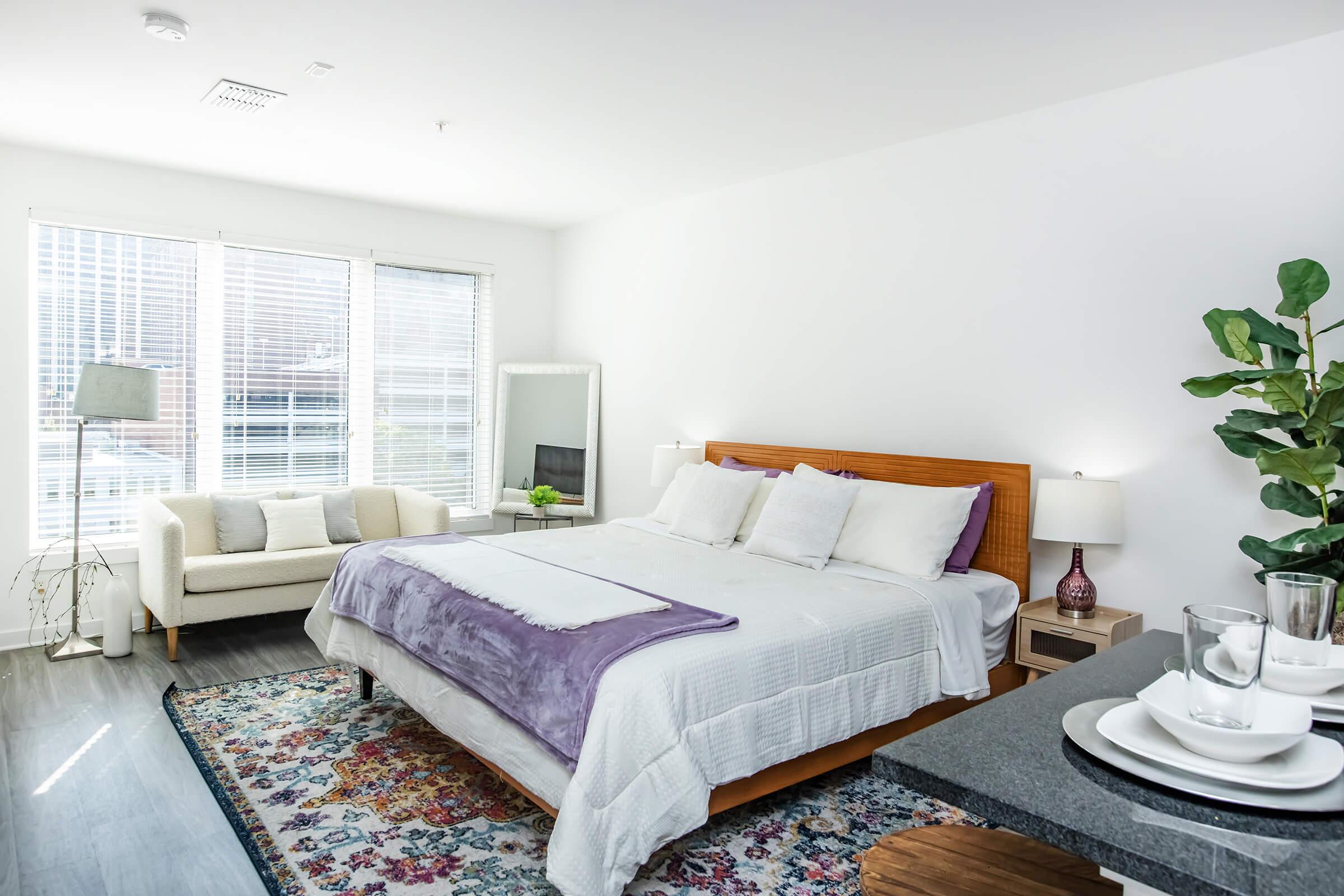 A bright and modern bedroom featuring a large bed with white linens and a purple throw. Natural light streams through large windows with blinds. There's a cozy seating area with a sofa and a minimalist lamp, as well as a small table set with plates and glasses, creating an inviting atmosphere.
