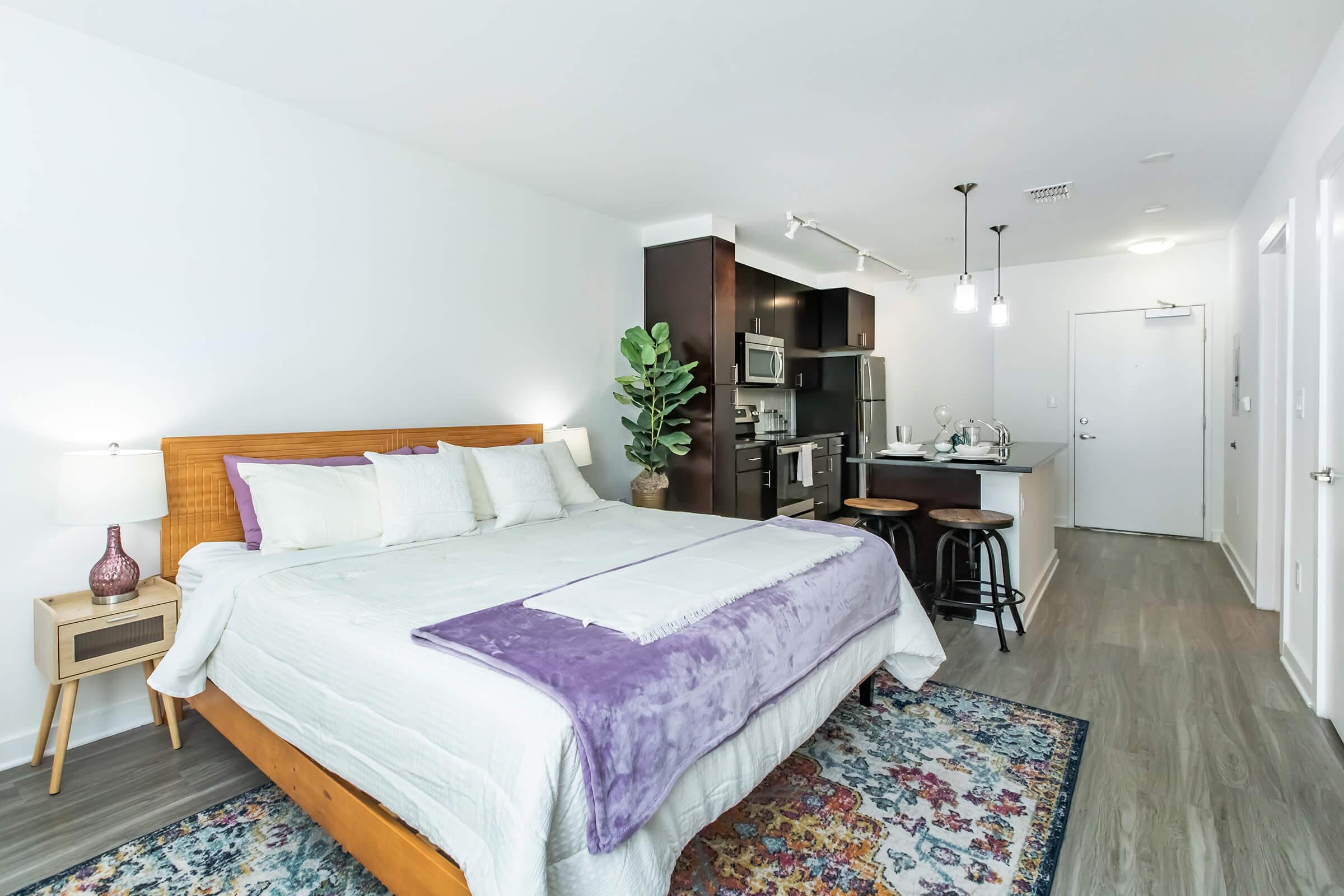 A modern studio apartment featuring a queen-size bed with white and purple bedding, a small kitchen area with a bar counter and stools, and a potted plant. The room has light-colored walls and wooden flooring, creating a bright and inviting atmosphere.